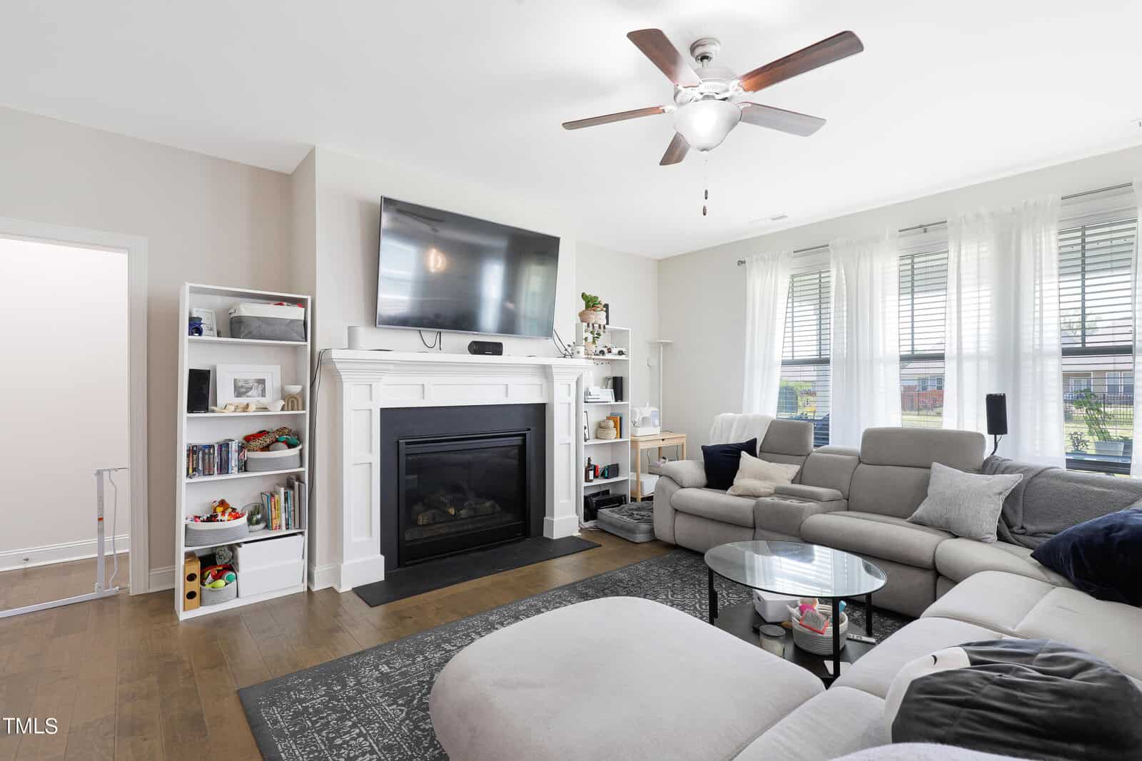 A cozy living room features a large sectional sofa with cushions, a mounted flatscreen TV above a fireplace, and a ceiling fan. Natural light streams through curtained windows, and shelves filled with books and decor flank the fireplace.