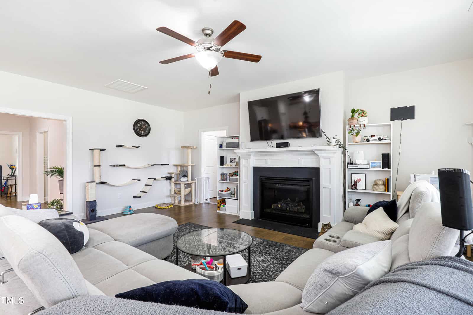 A cozy living room features a large L-shaped sofa, a ceiling fan, and a fireplace with a TV above it. Shelves adorned with various items are placed on either side of the fireplace, and a tall cat tree is in the corner.