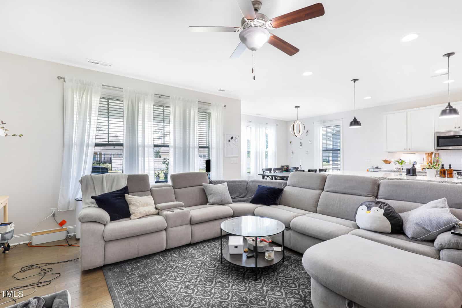 A bright living room with a large gray sectional sofa and a glass coffee table. A cute penguin cushion sits on the sofa. The room is well-lit with large windows, white curtains, and modern ceiling fan above. The open kitchen is visible in the background.