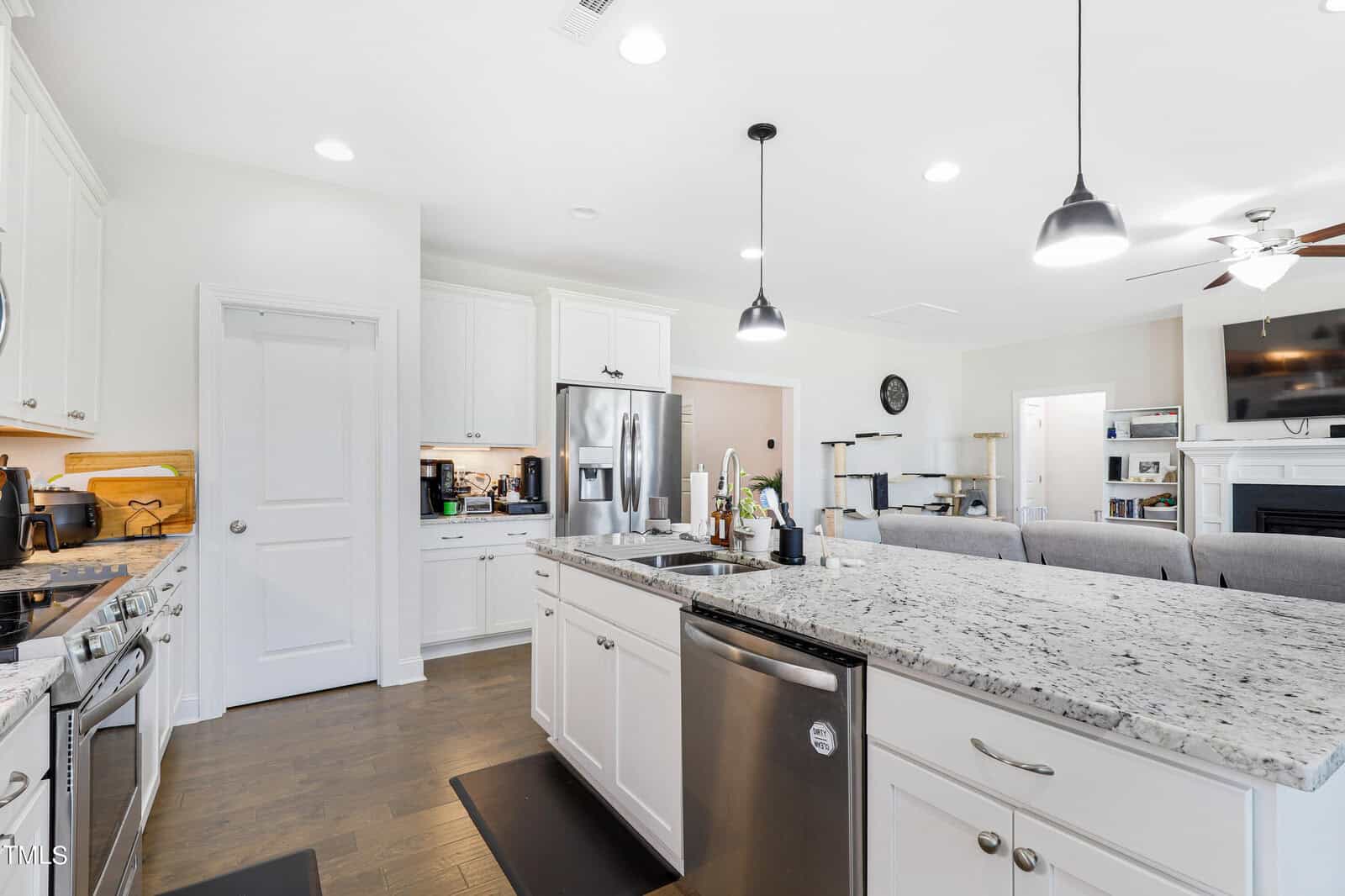 A modern kitchen with white cabinets, a gray granite island, stainless steel appliances, and dark wood flooring. Pendant lights hang above the island. The adjacent living room features a ceiling fan, large windows, and a visible flat-screen TV.