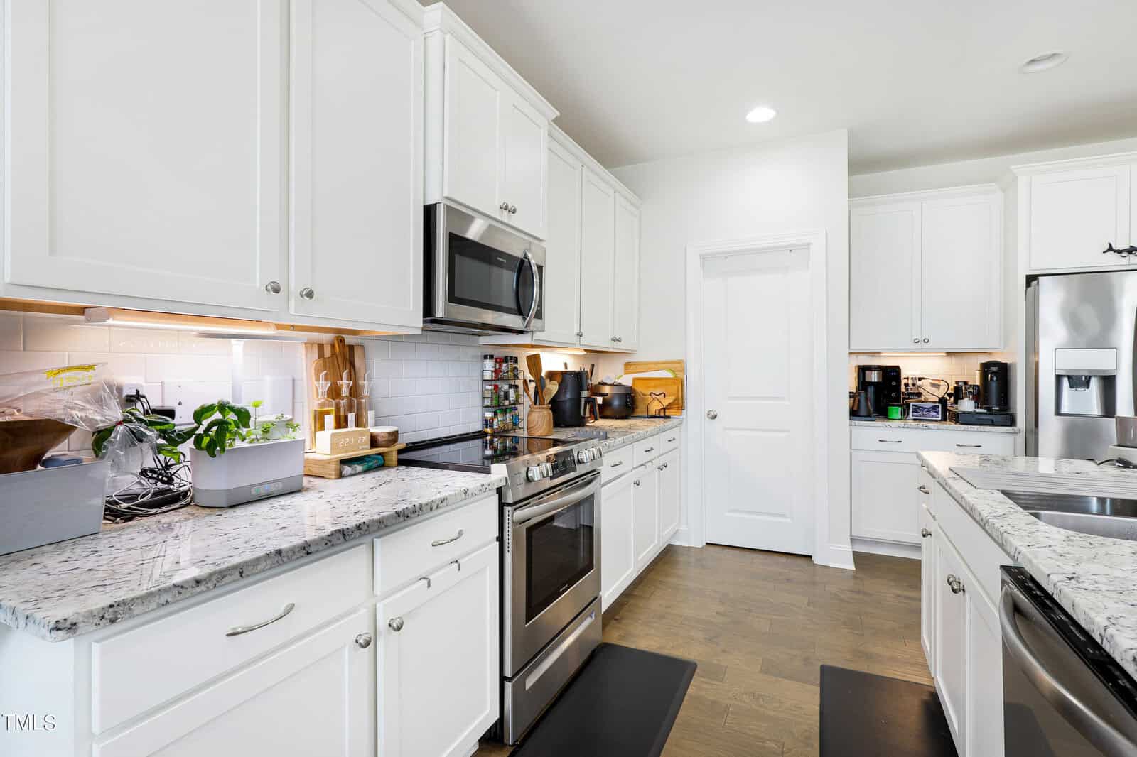 A modern kitchen featuring white cabinets, stainless steel appliances, and granite countertops. There are two black mats on the wooden floor, a plant near the backsplash, and a closed white door in the background.