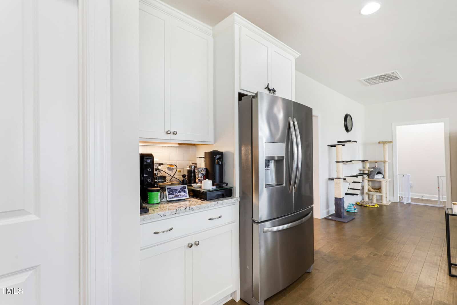 A modern kitchen featuring white cabinets, a stainless steel refrigerator, and a small countertop with a coffee maker and various items. In the background, a cat tree, a toy, and a clock on the wall are visible on the wooden floor.