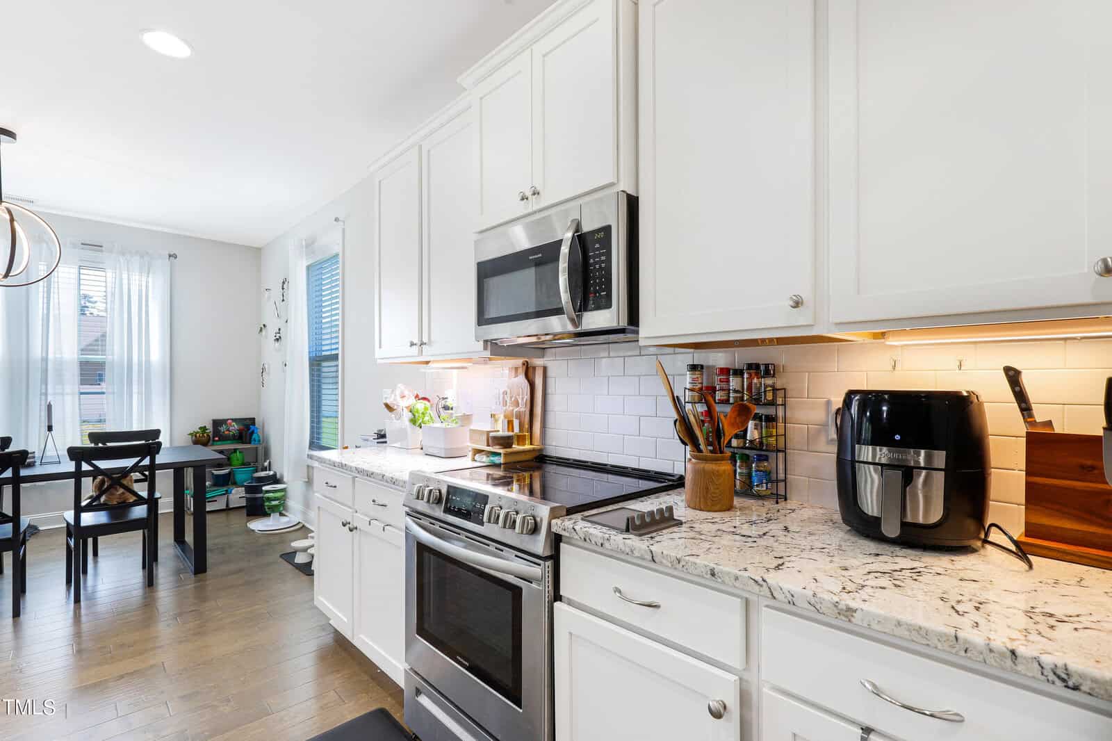 A modern kitchen with white cabinets, a granite countertop, and stainless steel appliances, including a microwave and stove. An air fryer sits on the counter. The kitchen opens to a dining area with a black table and chairs near a window.