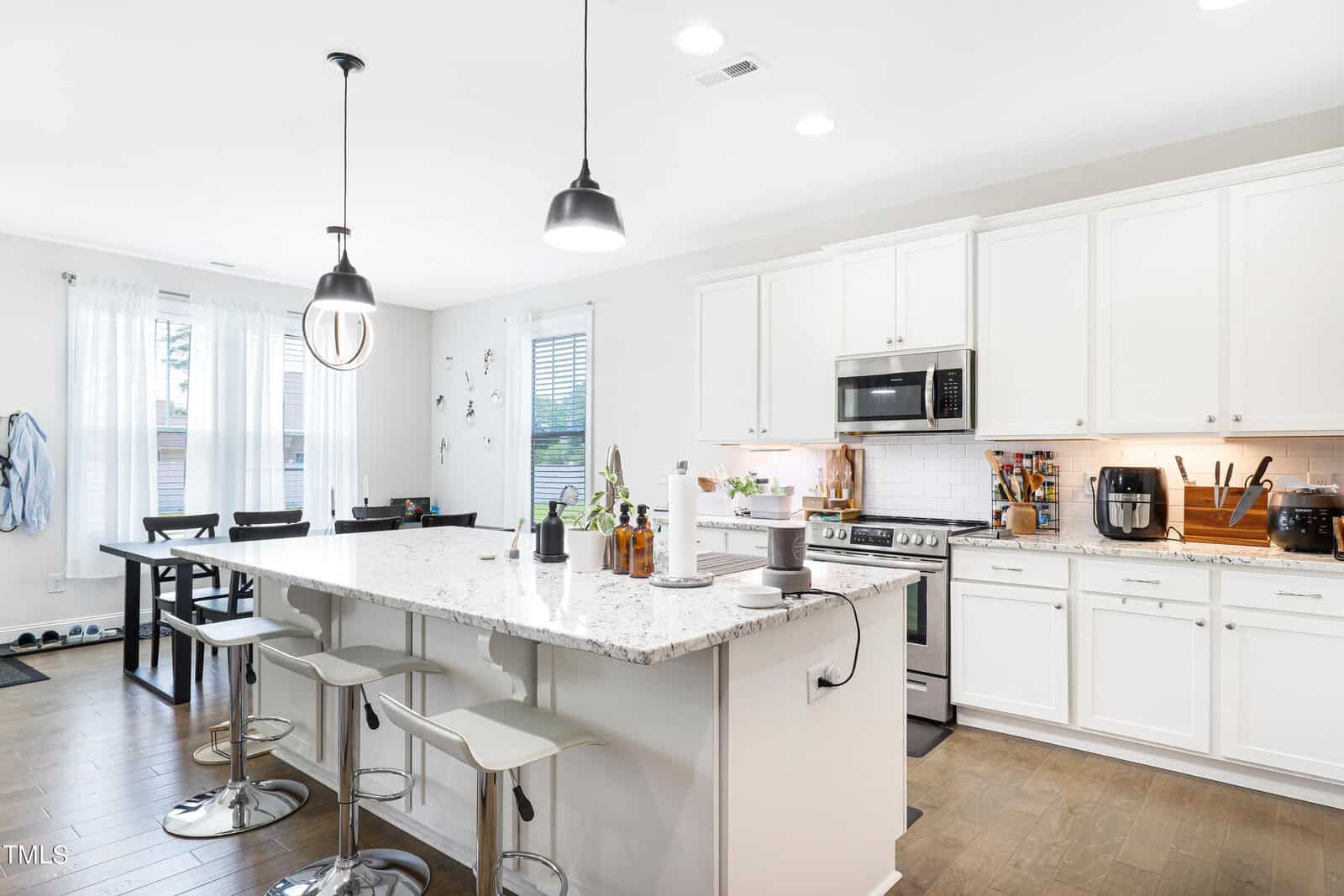 A modern kitchen with white cabinets and a marble island countertop. Three gray bar stools line the island, which is equipped with a blender and other kitchen items. Pendant lights hang above. A dining area with a table is visible in the background.