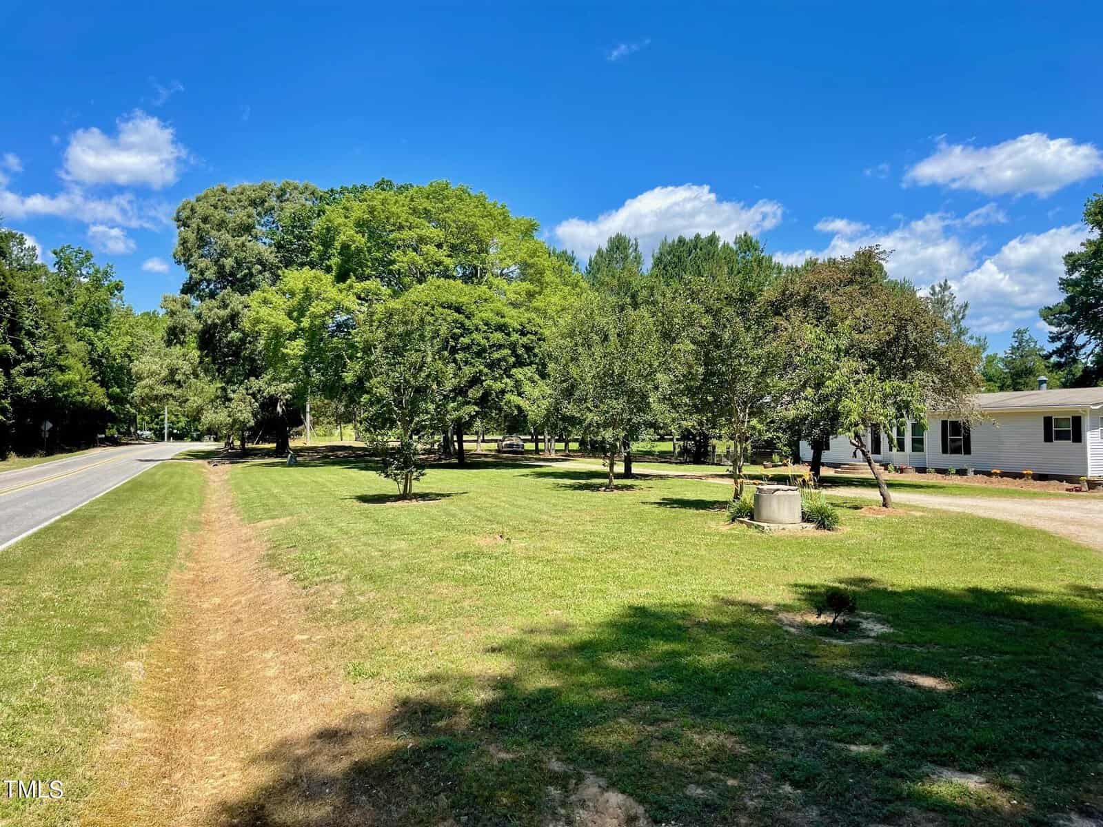 A sunny rural scene with a grassy lawn, several trees, and a white house on the right. A two-lane road runs adjacent to the lawn on the left. A small well-like structure is visible near the house. The sky is clear with a few scattered clouds.