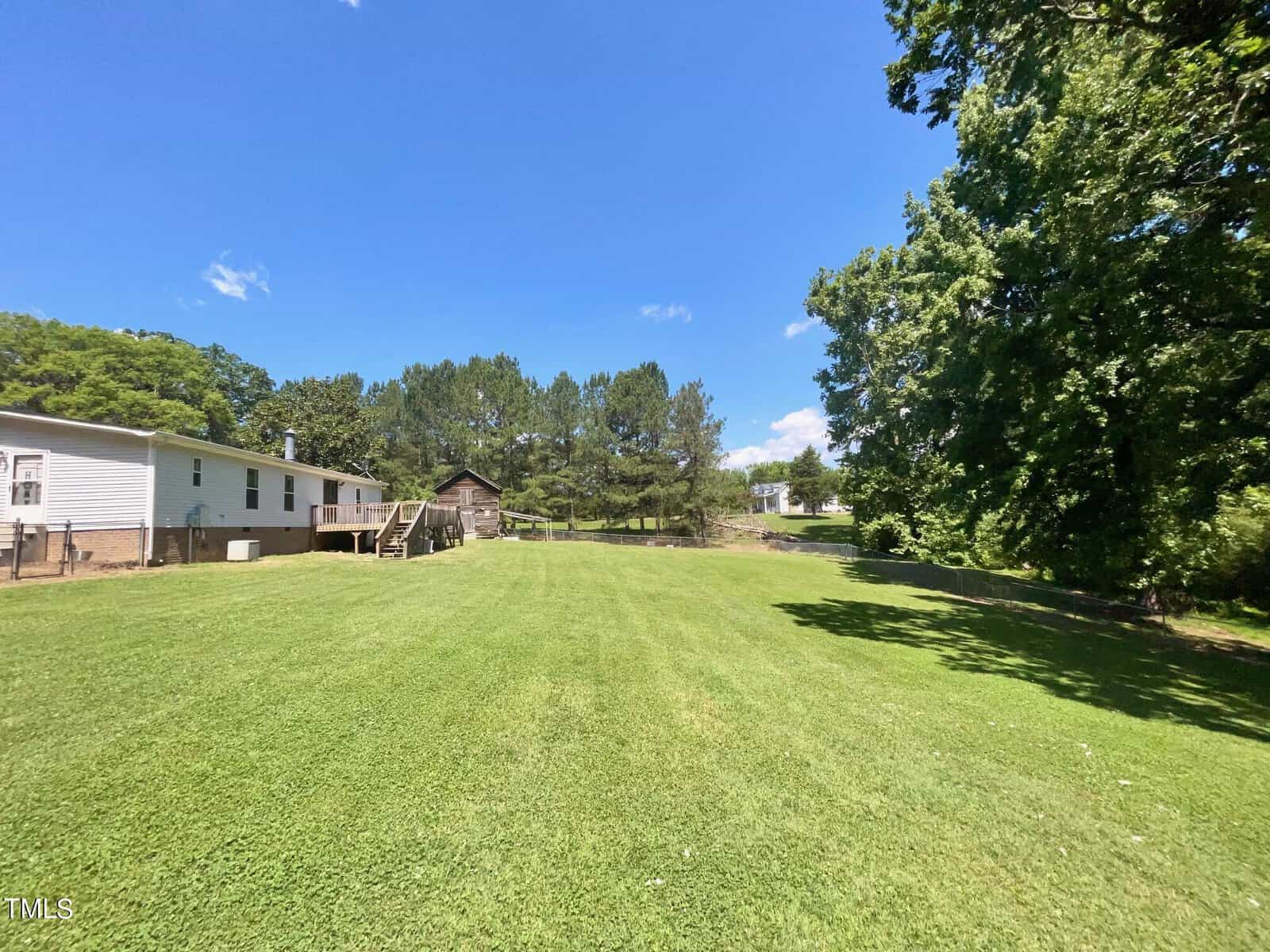 A spacious, well-kept backyard with a large green lawn stretches out under a clear blue sky. A house with an elevated deck is visible on the left side, and several mature trees frame the yard, providing a mixture of open space and shaded areas.