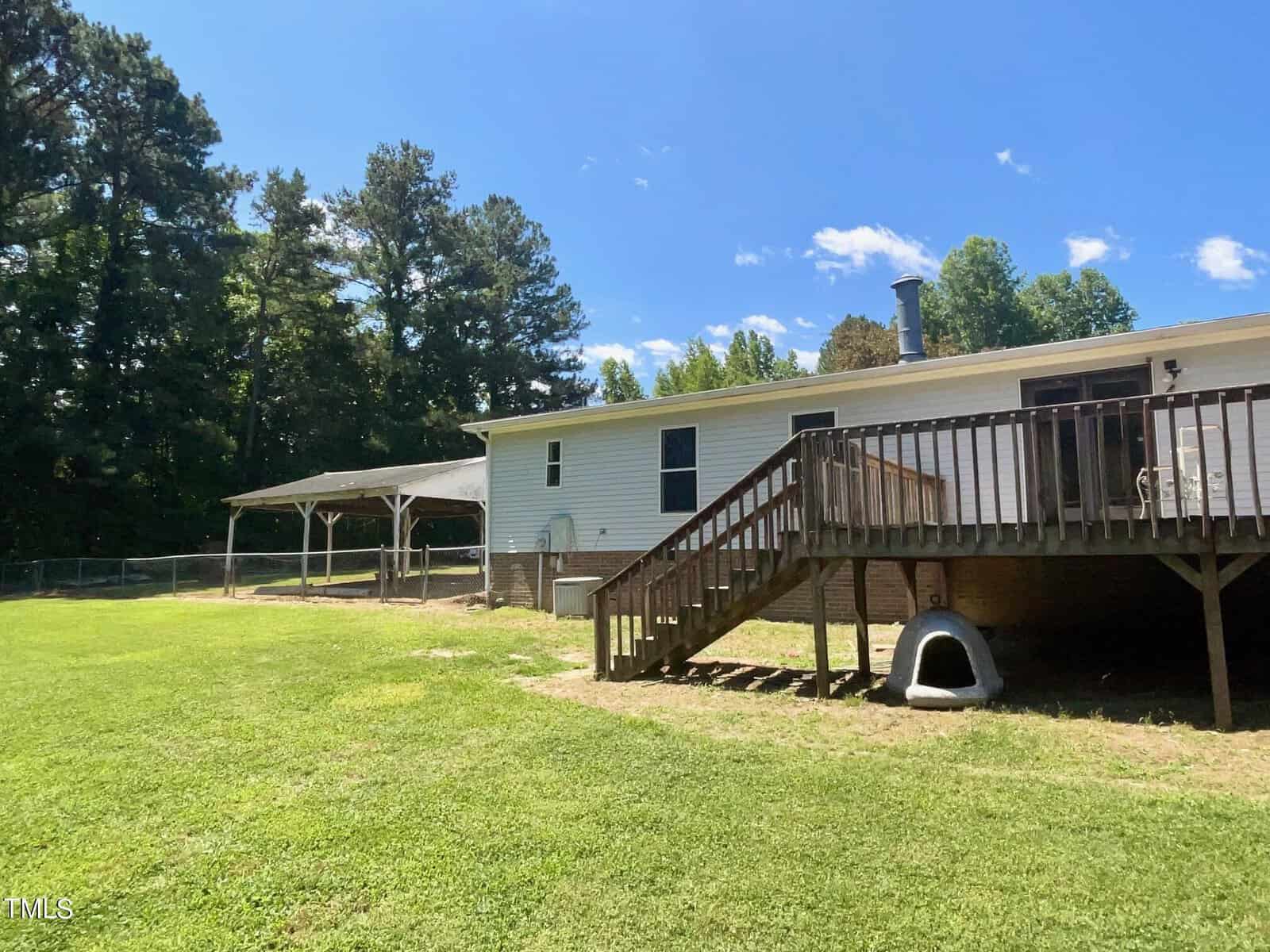 A backyard scene features a white single-story house with a raised wooden deck, accessible by a set of stairs. A dog house is placed underneath the deck. The yard includes a grassy area and a canopy-covered structure on the left side. Tall trees line the background.