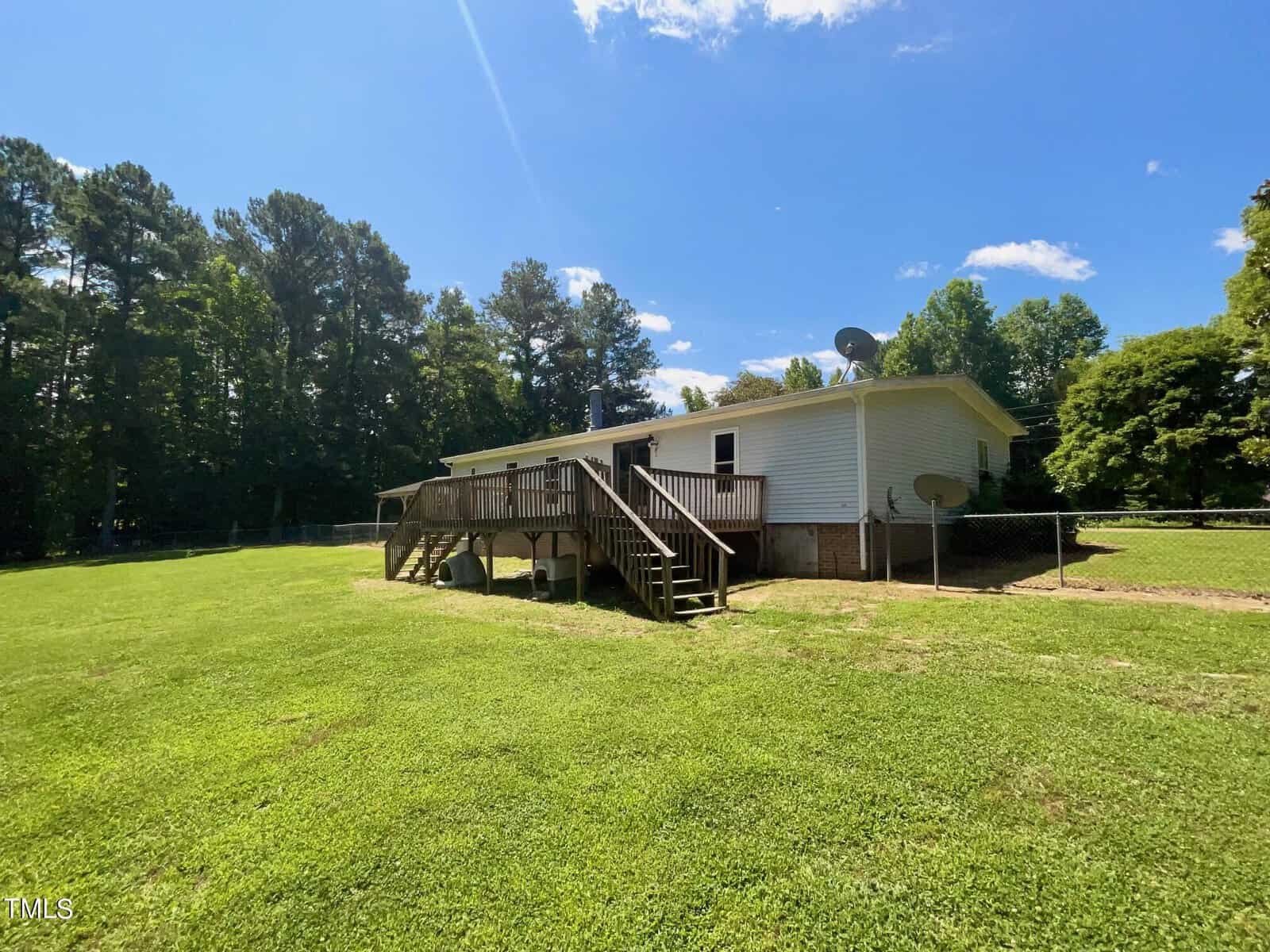 A white, single-story house with a wooden deck and stairs leading to the backyard. The property is surrounded by a grassy lawn and tall trees in the background. Two satellite dishes are attached to the side of the house. The sky is clear and sunny.