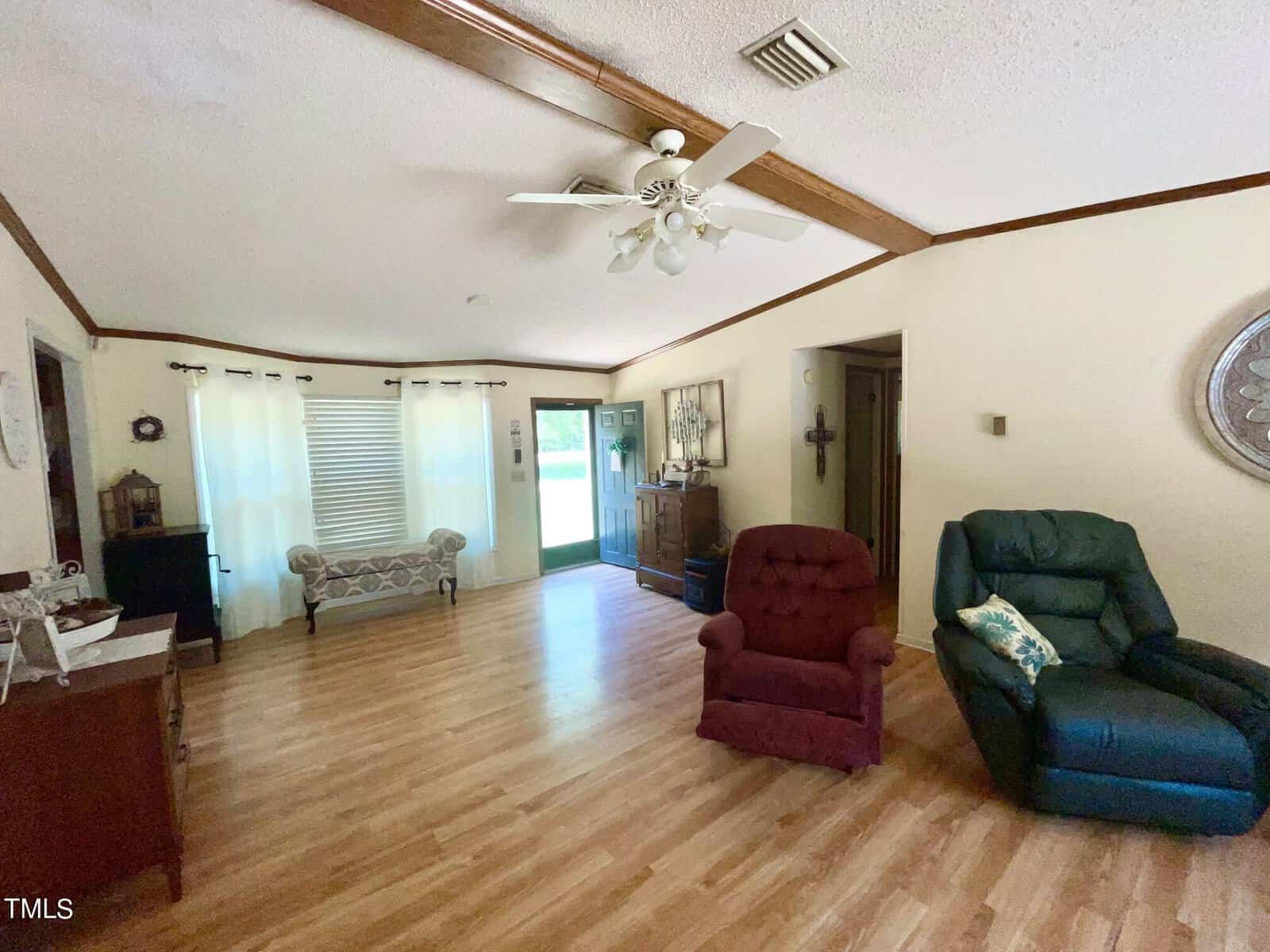A spacious living room with hardwood floors and a vaulted ceiling with exposed beams, featuring a green recliner, red armchair, and a long cushioned bench. A ceiling fan is mounted above, and two sets of curtained windows allow natural light to brighten the room.