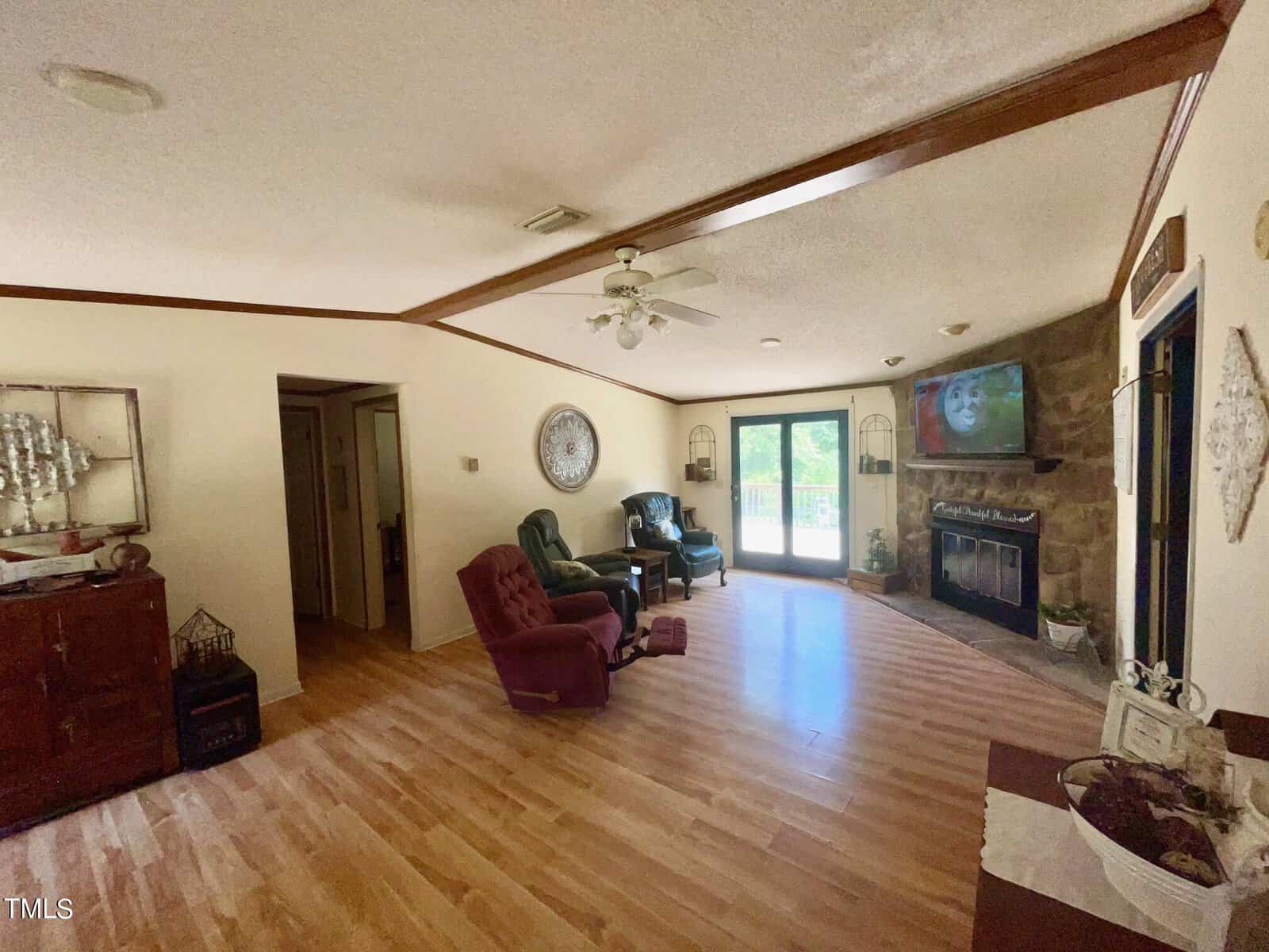 A cozy living room with wood flooring, a stone fireplace, and a flat-screen TV mounted above it. Two armchairs, one purple and one green, are arranged around the fireplace. Sliding glass doors lead to an outdoor area. Miscellaneous decorations and a ceiling fan are visible.