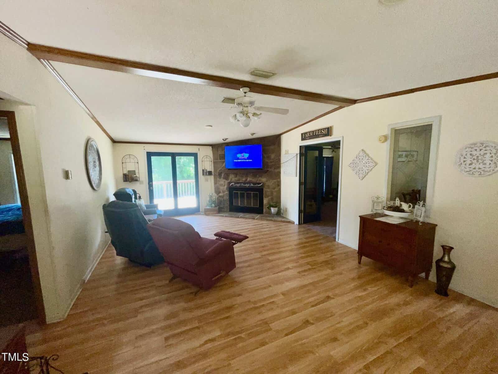 A living room with light-colored hardwood floors, a recliner, a TV mounted above a fireplace, and glass doors leading to a patio. Decor includes a ceiling fan, a wooden dresser, and wall art. Doorways lead to other rooms. Natural light enters through the glass doors.
