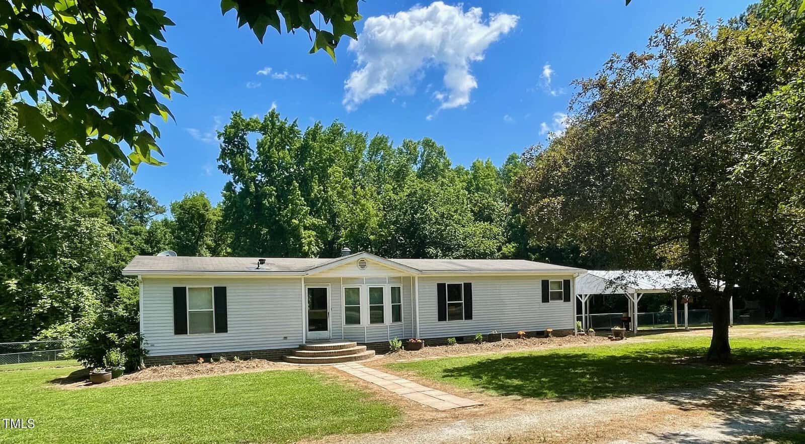 A white, single-story house at 3520 Egypt Mountain Road, Kittrell, with a light grey roof is surrounded by lush green trees under a bright blue sky with a few clouds. There is a small garden in front, a path leading to the entrance, and a carport on the right side. The lawn is well-kept.