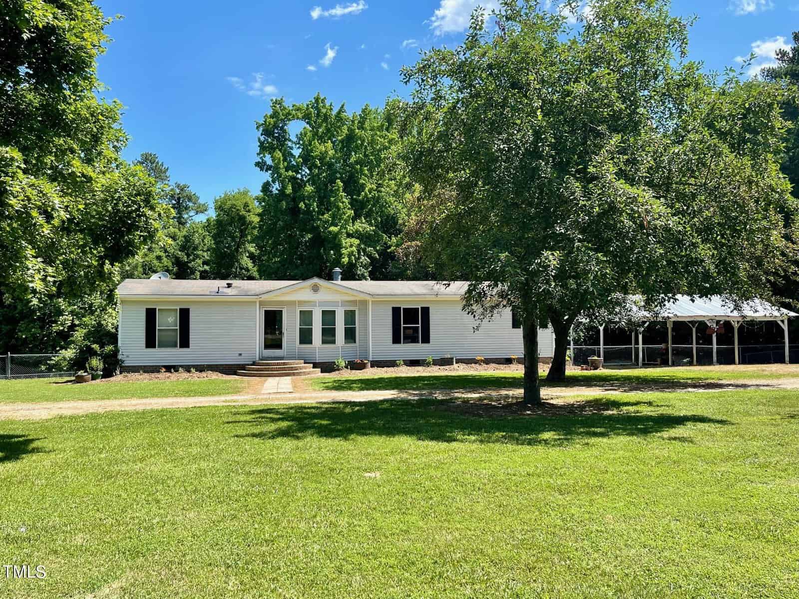 A single-story white house with dark shutters sits amid a lush green lawn. A large tree stands in front, and a gravel path leads to the entrance. To the right, there's a carport. Trees and shrubbery form a dense backdrop against a clear blue sky.