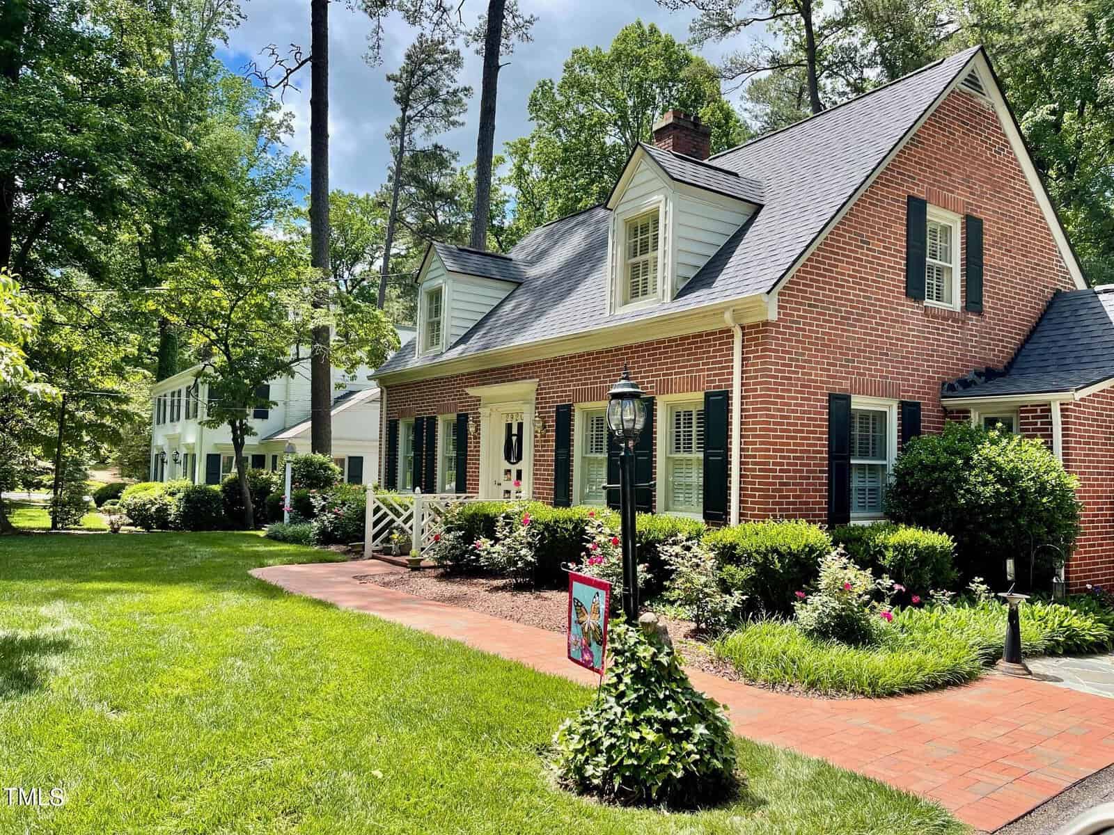 A charming brick house at 2920 Dogwood Drive in Henderson, NC, with a black roof, white shutters, and a white front door. The home features a well-manicured lawn, green shrubs, and a flower bed along the brick walkway leading to the entrance. Tall trees surround the property, providing shade and a peaceful atmosphere.