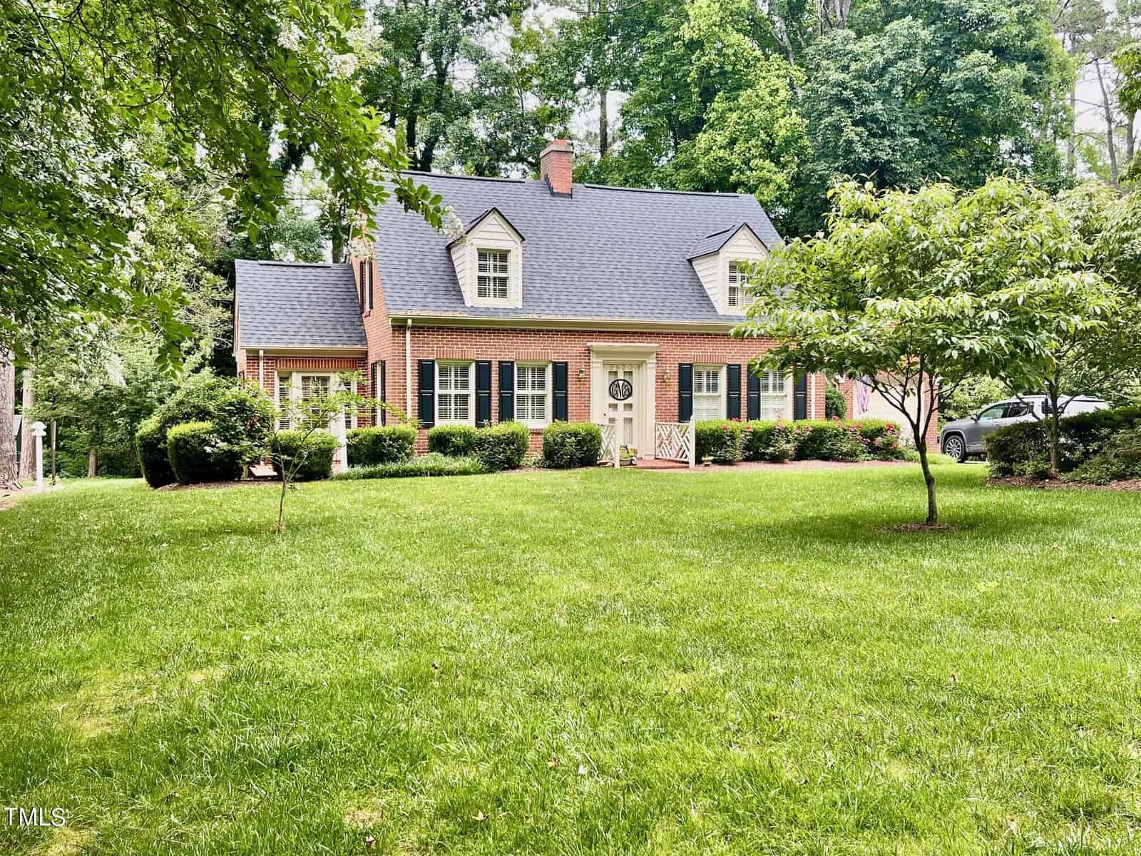 A charming brick house with two dormer windows and a central door, surrounded by lush green grass and trees. The house has a well-maintained front yard with bushes and a small tree to the right. A car is partially visible on the driveway on the right side.