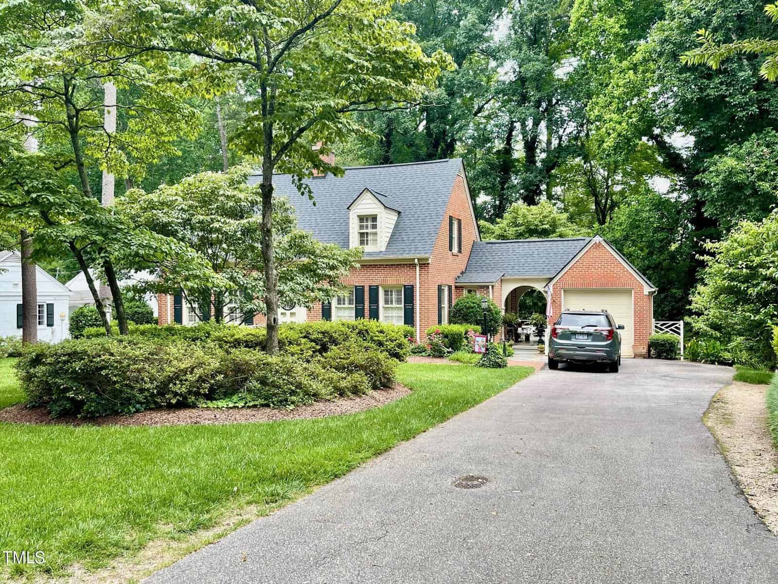 A red-brick house with white trim and black shutters has a well-maintained garden featuring green shrubs and trees. A grey car is parked in the driveway, partially under a carport attached to the house. The background consists of tall, leafy trees.