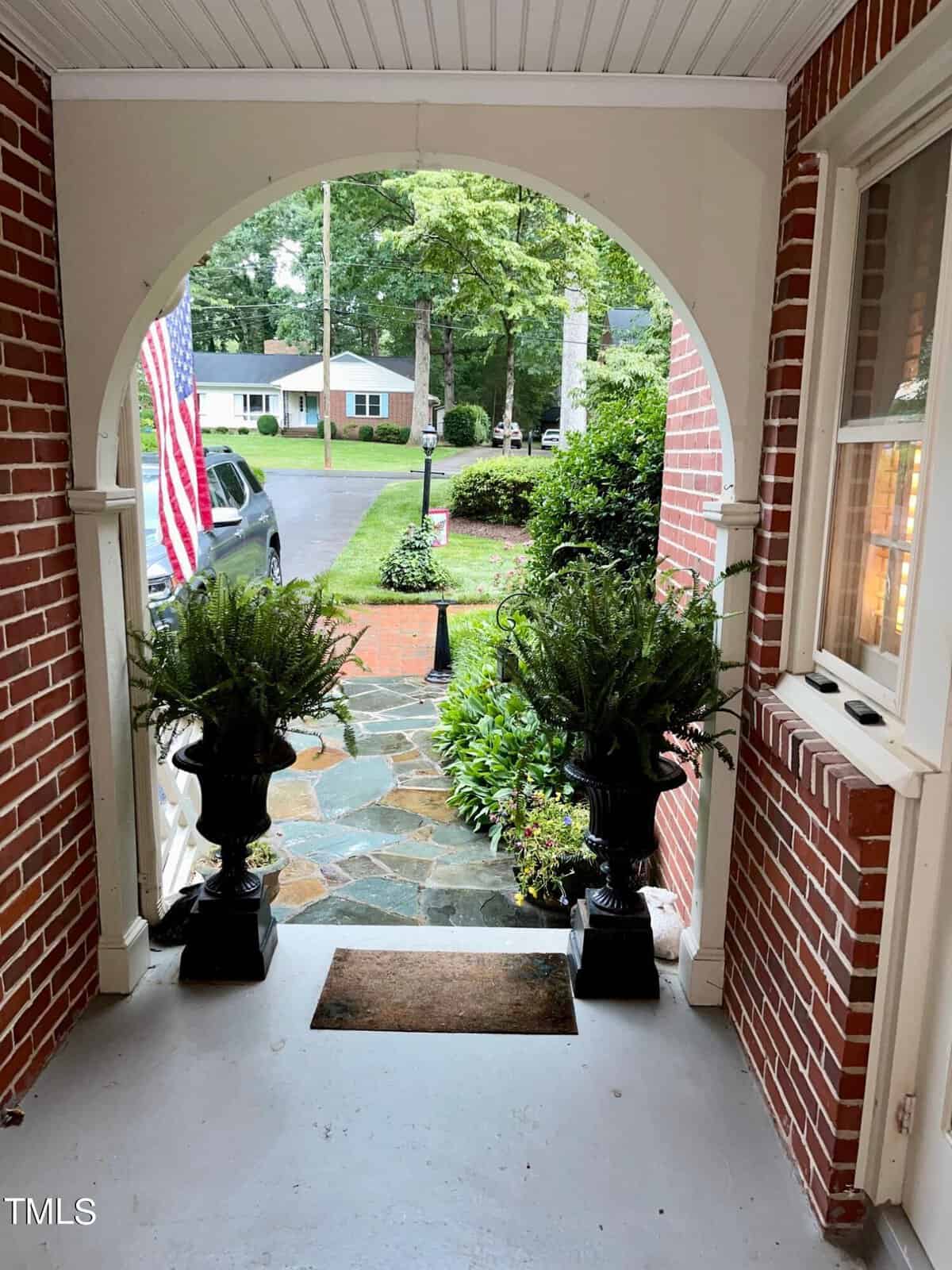 A brick archway frames a porch and pathway leading to a well-maintained garden and a suburban street. Two large potted ferns sit on either side of the porch entrance. An American flag is displayed on the left side of the garden. A lawn and houses are visible.