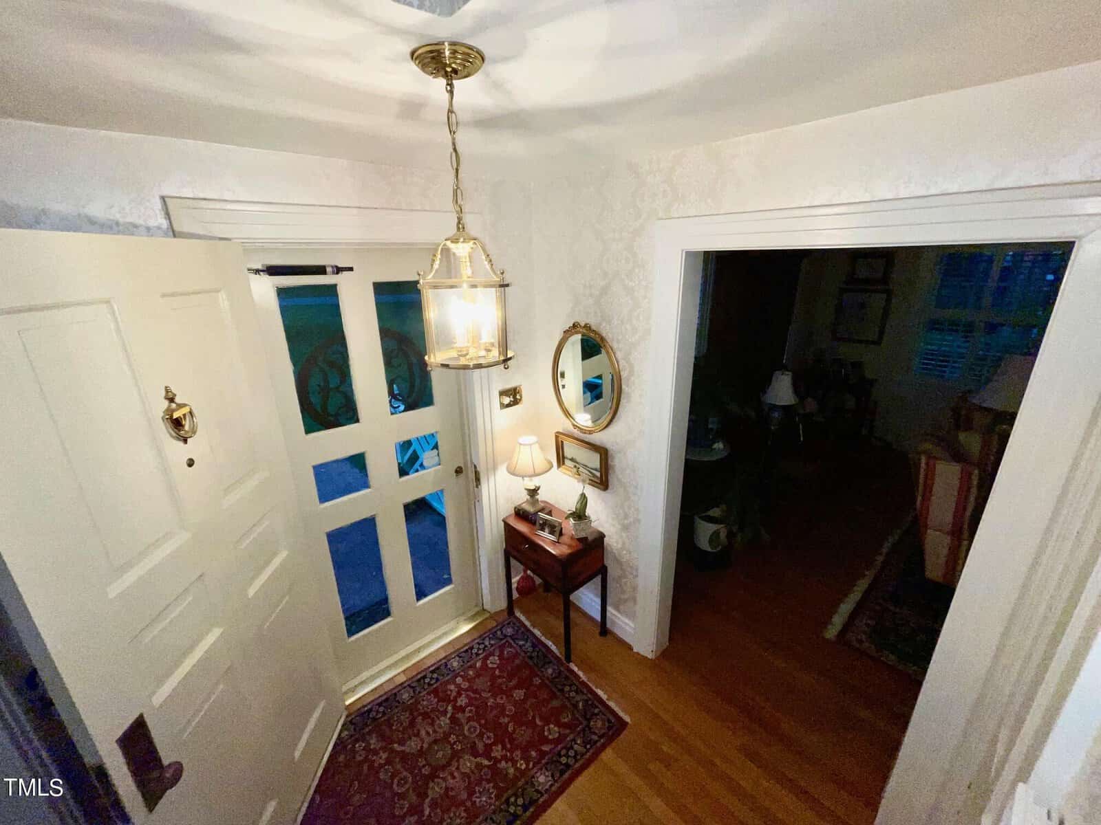 A warmly lit, entryway of a home features a white door with glass panels, a hanging pendant light, a small wooden table with a lamp and framed pictures, a round mirror above it, and a patterned red rug on wooden floors. Adjacent room is partially visible.