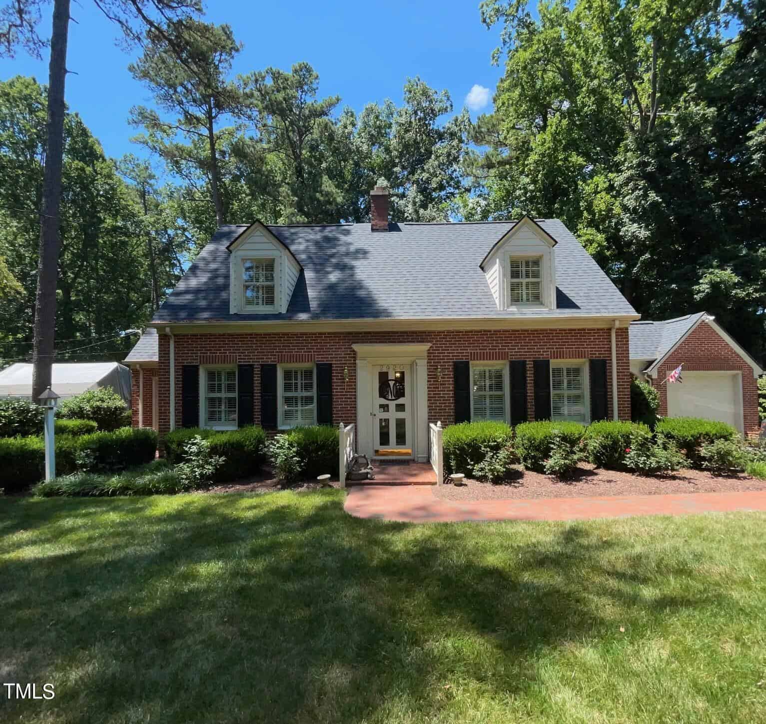 A charming red-brick house with a dark gray roof and two dormer windows. The entrance features a small porch with white railings and steps flanked by well-manicured bushes. White-trimmed windows with black shutters line the front. Tall trees and lush greenery surround the property.