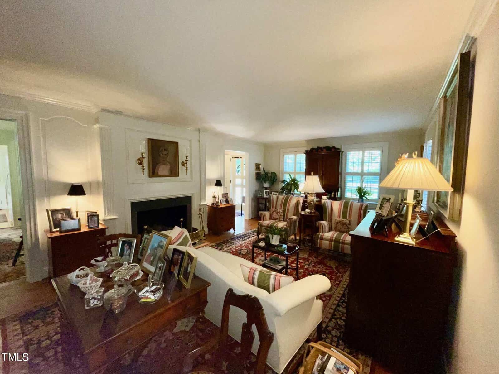 A cozy, traditional living room features a white fireplace, various framed photographs on a wooden table, a cream-colored sofa, and two armchairs with a glass coffee table in between. The room is warmly lit by table lamps and has large windows with green plants.