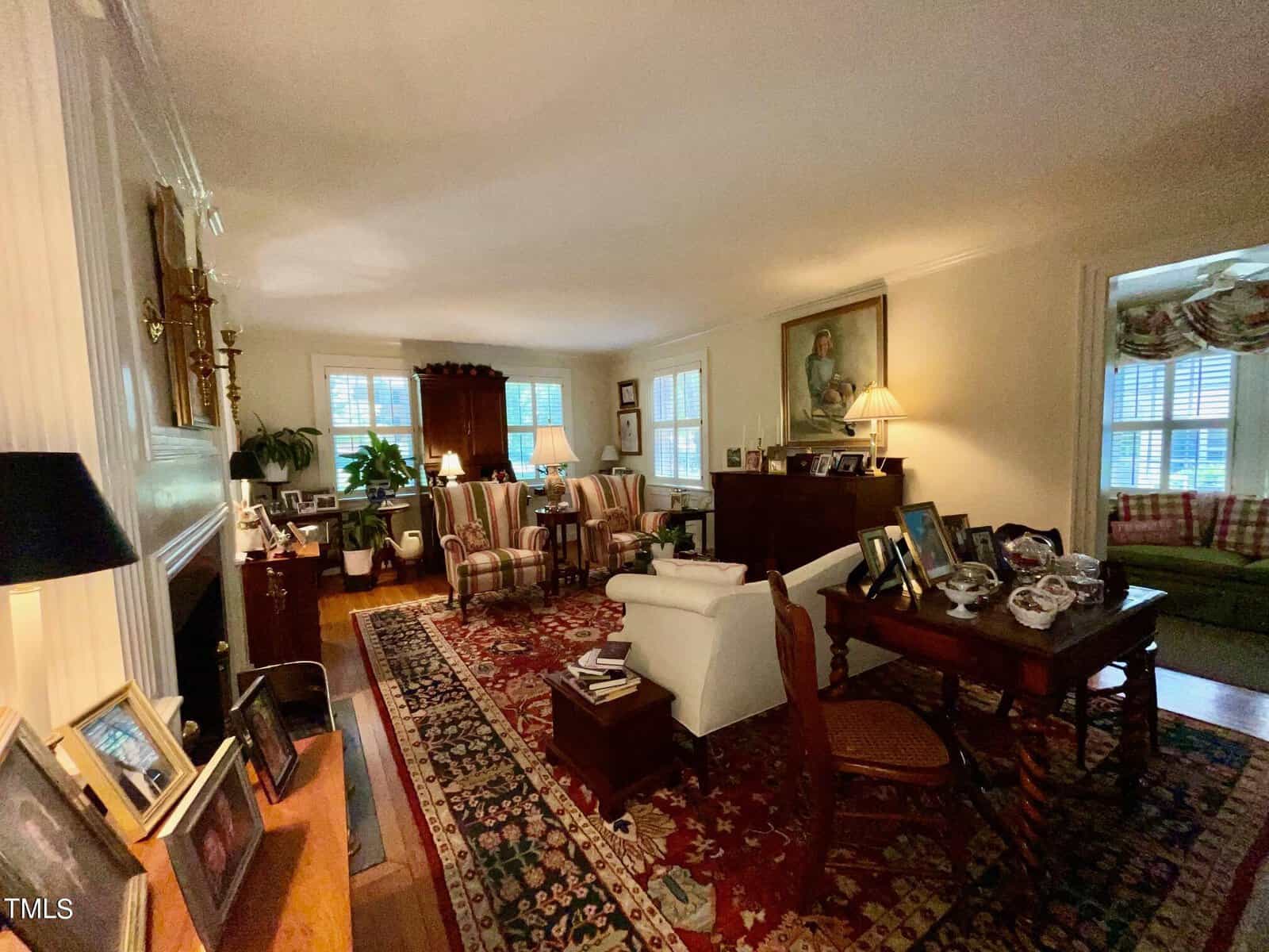 A cozy living room with traditional decor, featuring a dark wood cabinet, white sofa, armchairs, and a wooden table with framed photos and decorative items. Large windows allow natural light in, and a colorful patterned rug covers the hardwood floor.