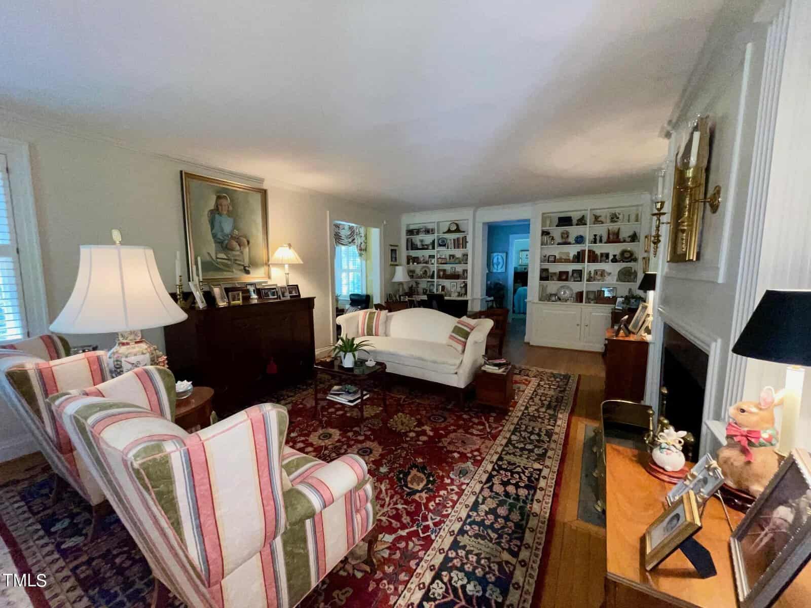 A cozy living room with patterned armchairs, a white sofa, and a large rug. Shelves filled with family photos line the walls, and the room is decorated with lamps, a fireplace, and art. The space has a warm, inviting atmosphere with a mix of vintage and modern elements.