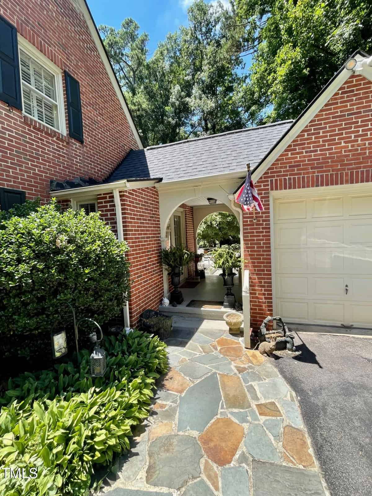 A charming brick house with a stone-paved pathway leading to an arched entrance adorned with plants. An American flag hangs from the porch near the garage. Bushes and trees surround the house, creating a lush, green environment.