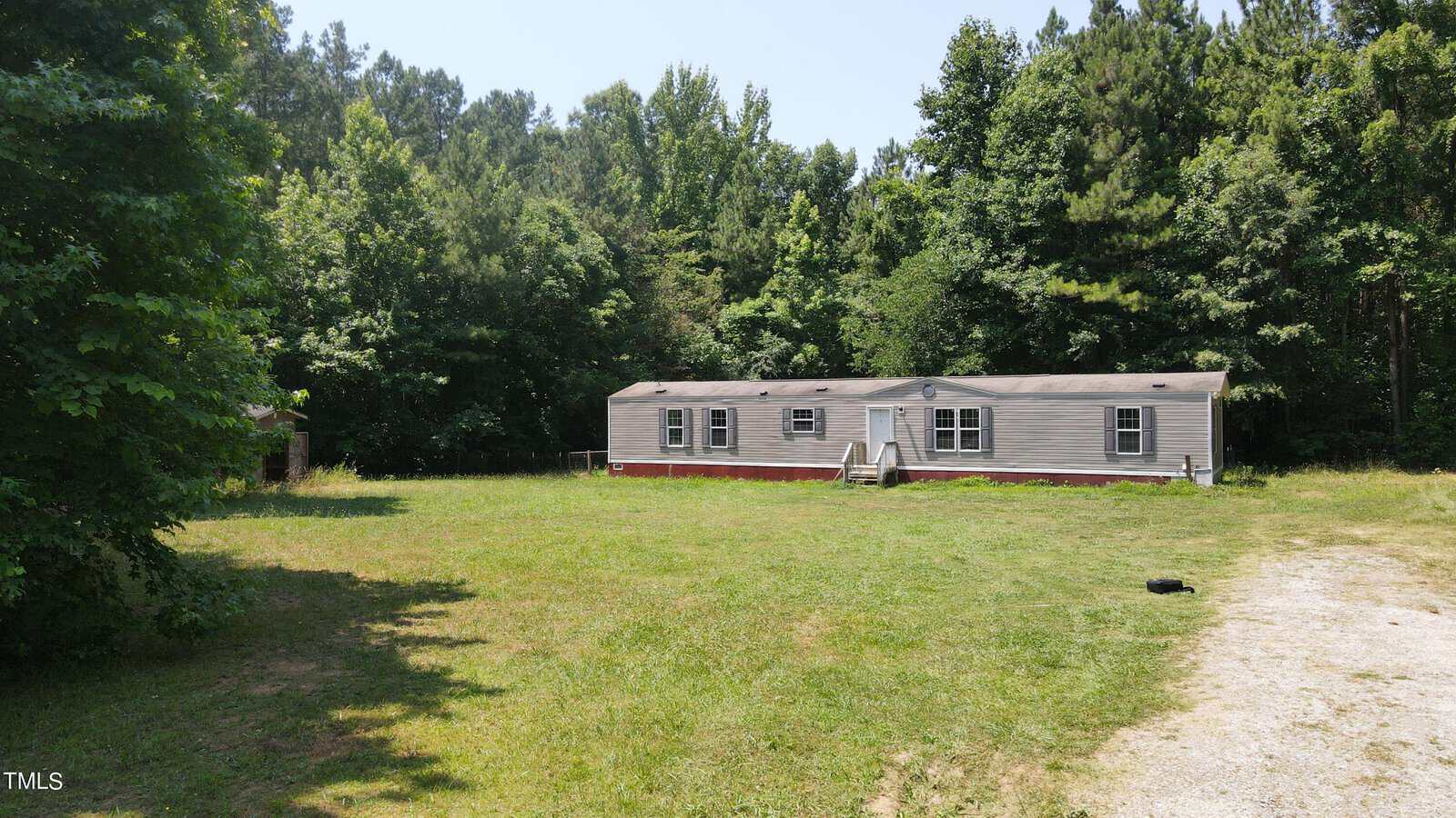 A single-story mobile home sits on a grassy plot surrounded by trees. The home is light gray with a small set of steps leading to the front door. A gravel path is visible in the foreground, curving to the right.