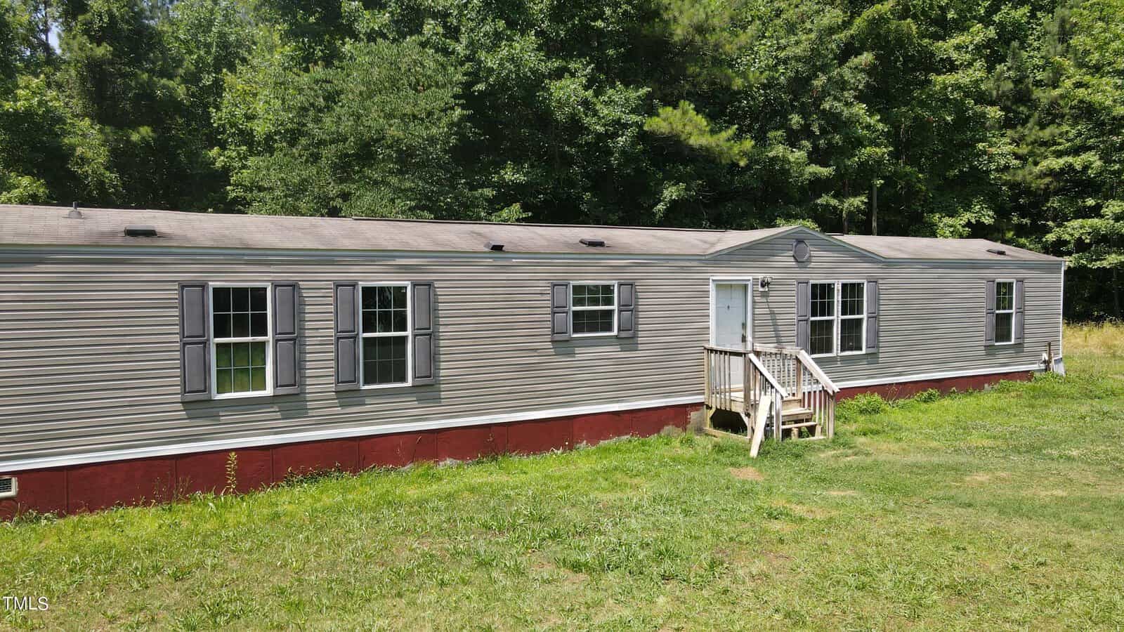 A long, light gray mobile home with a red foundation is set on a grassy lawn, surrounded by dense green trees. The home features several windows and a small wooden staircase leading to the entrance.