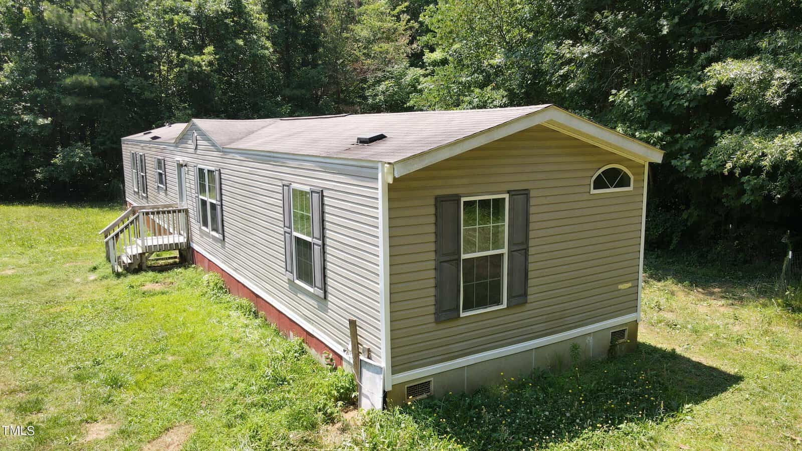 A beige mobile home with brown shutters is situated on a grassy lawn, bordered by dense, green foliage. A small wooden staircase leads to the entrance on the side. The roof has two vent openings, and a clear blue sky is visible above.