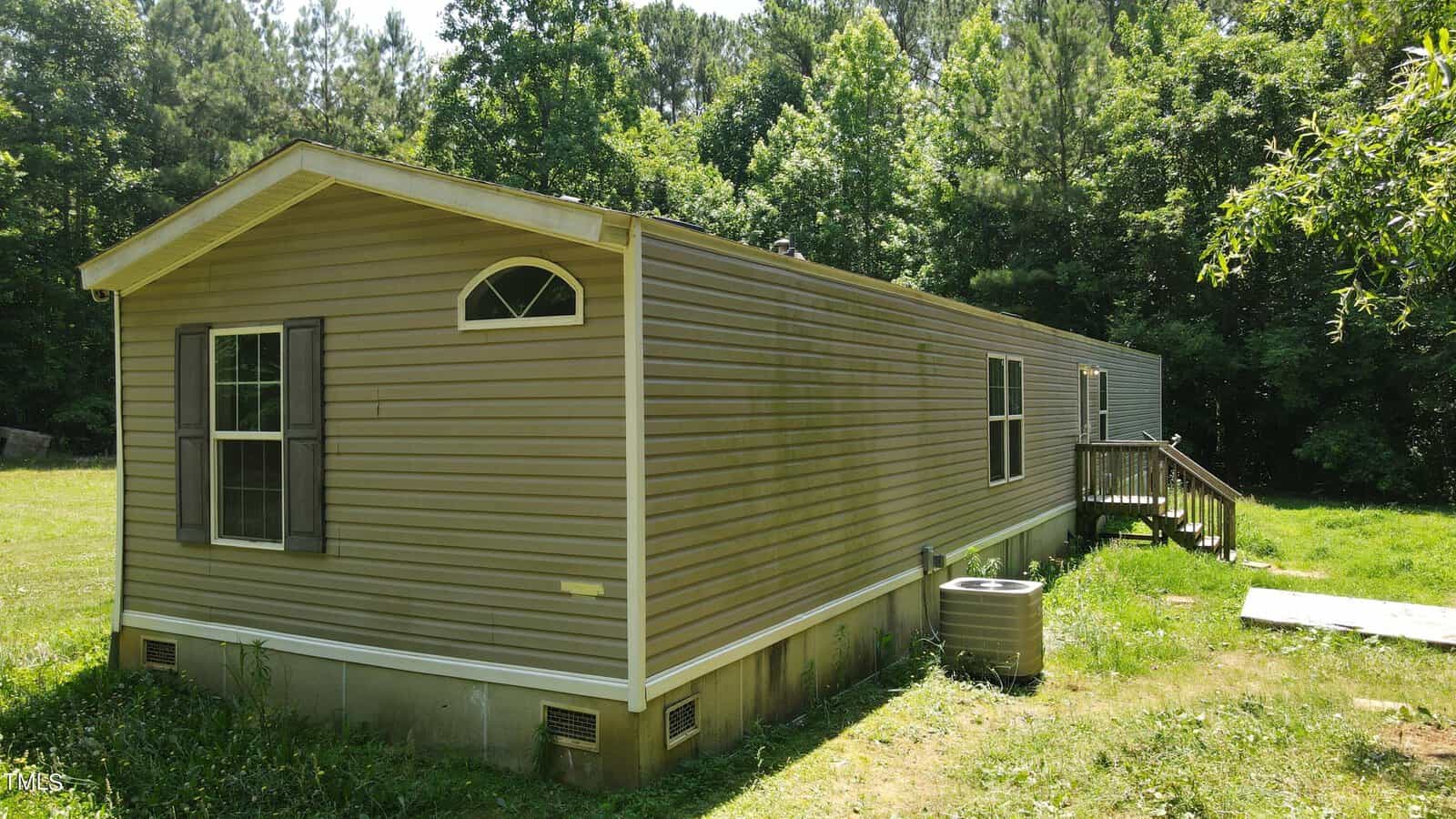 A beige mobile home with a small wooden staircase leading to the entrance, surrounded by grass and trees in the background. The home has a single window with shutters and an arched window near the roof.