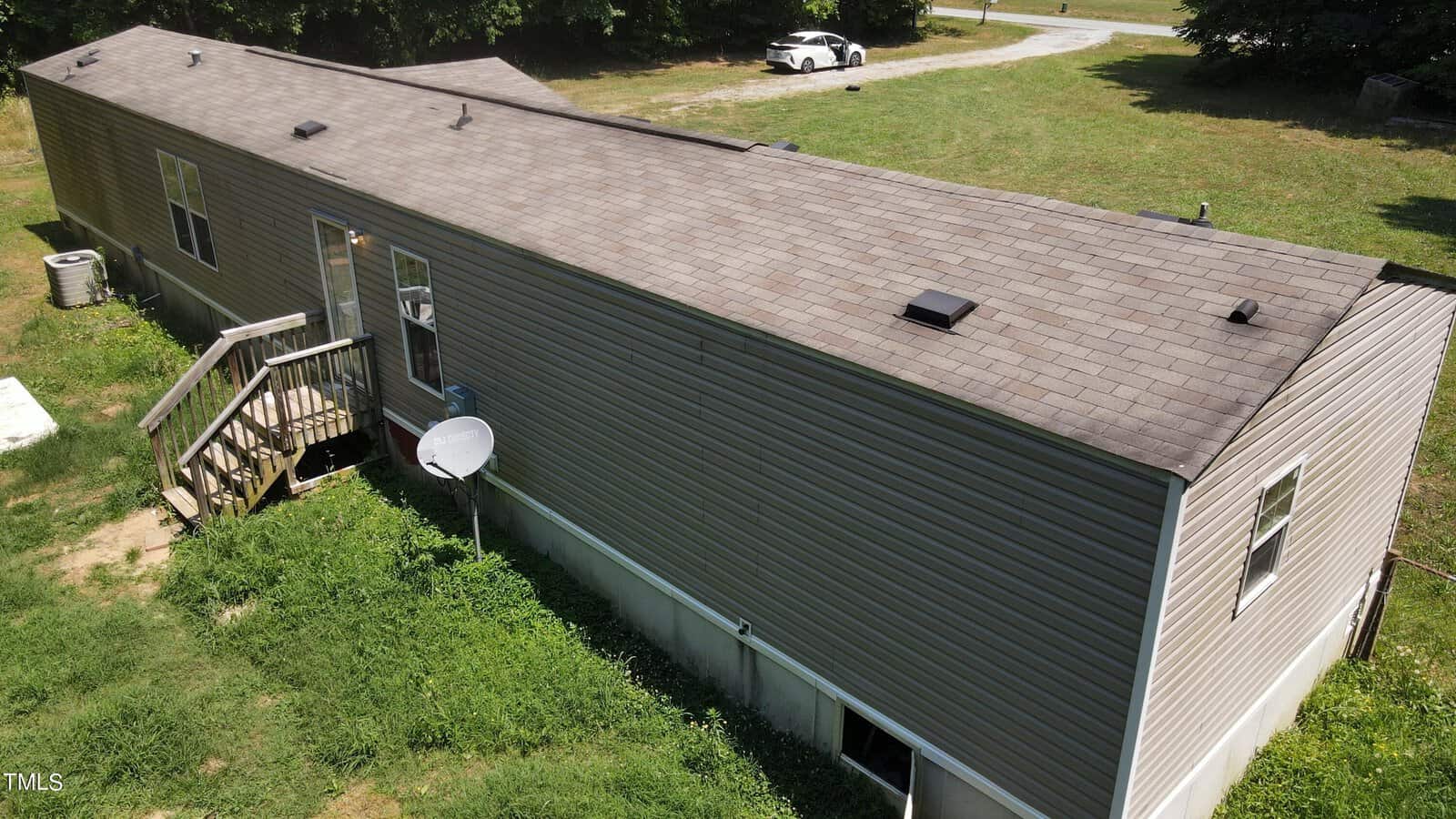 Aerial view of a single-story mobile home with a gray roof and siding, surrounded by grass. A small wooden staircase leads to the entrance. A satellite dish is attached near the stairs. A white vehicle is parked on a gravel path nearby.