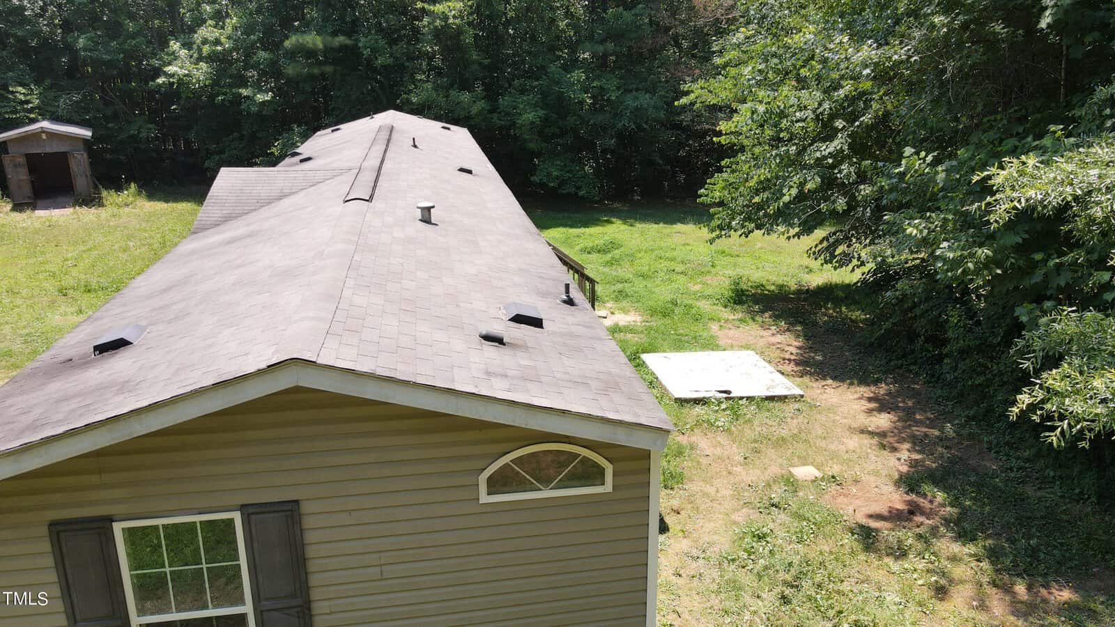 Aerial view of a long, single-story house with a gray roof surrounded by a grassy yard and dense green trees. A small shed is visible in the background. Sunlight casts shadows on the ground.