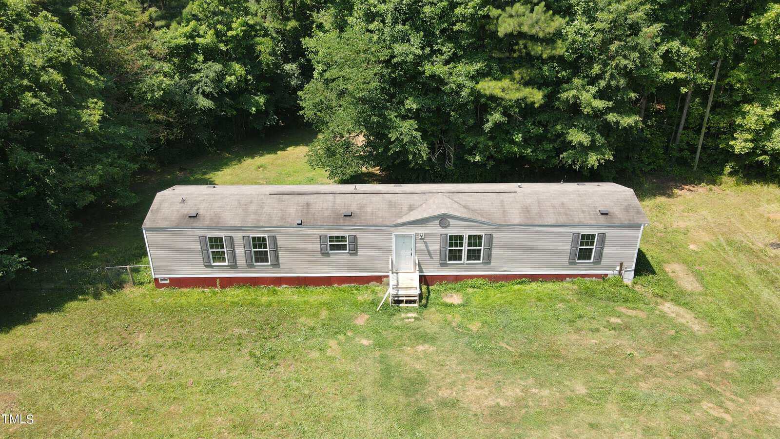 Aerial view of a single-story, rectangular mobile home with a light gray exterior and red foundation, surrounded by grass and backed by dense trees under a clear sky. Steps lead to the front door.