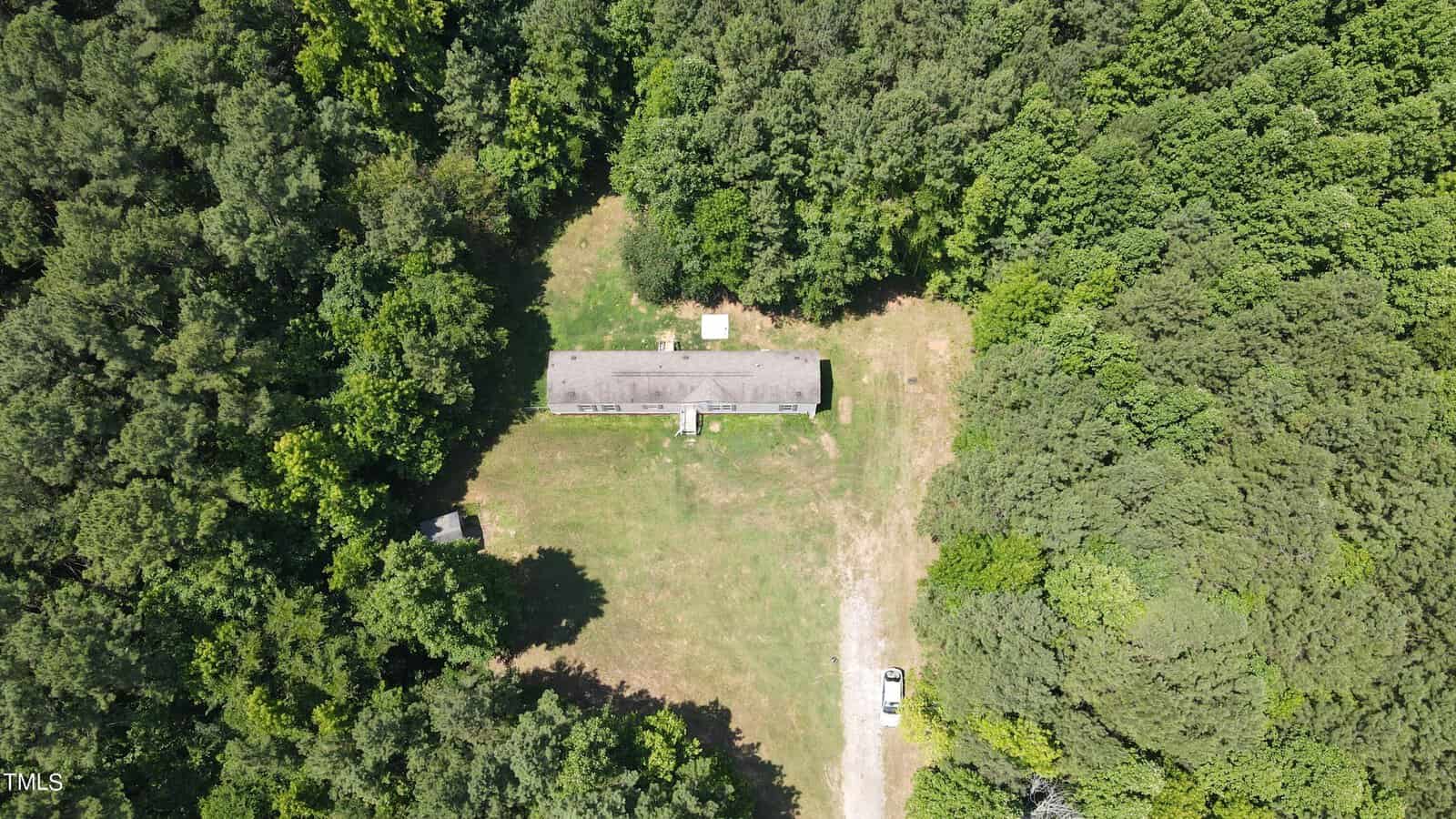 Aerial view of a single-story building surrounded by dense green forest. The clearing around the building features a grassy area and a parked white vehicle. A small path leads to the building, which has a rectangular layout.