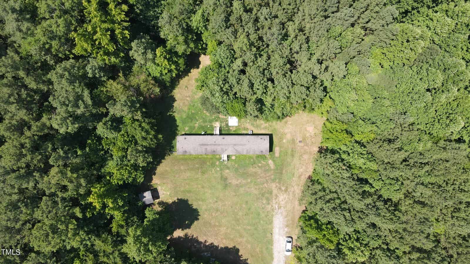 Aerial view of a long, narrow house surrounded by dense green forest. The house is centrally located in a clearing with a patch of grass and a vehicle parked nearby. A small structure is visible to the left of the house.