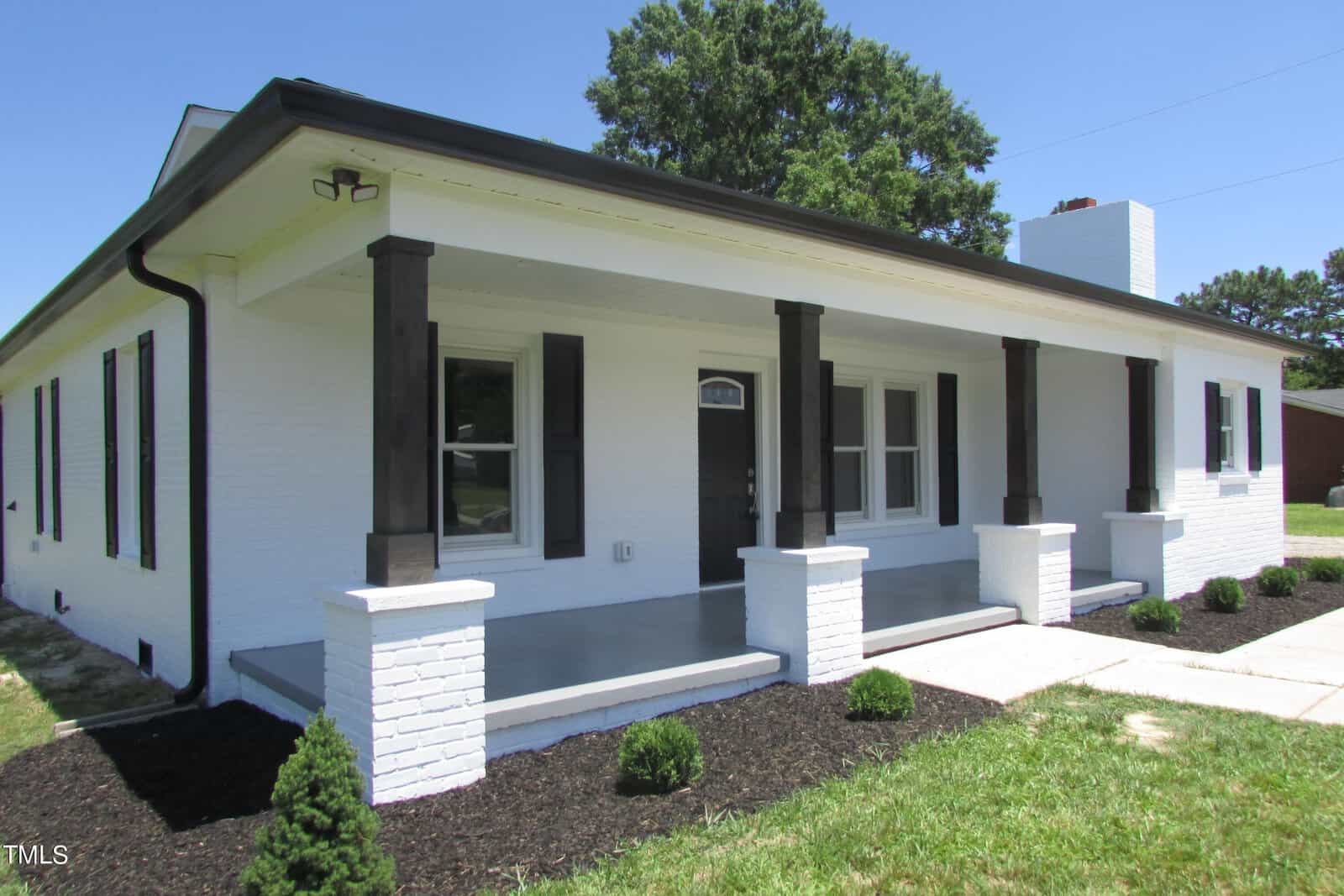 A white one-story house with a dark roof and dark trim, featuring a covered front porch with columns, a concrete walkway, and neatly landscaped bushes in front. Tall trees and a blue sky are visible in the background.