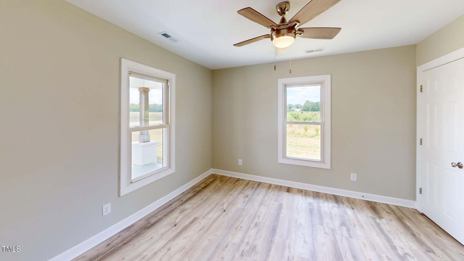 A small empty room with light wood flooring, beige walls, two windows, a white door, and a ceiling fan with wooden blades and a light fixture.