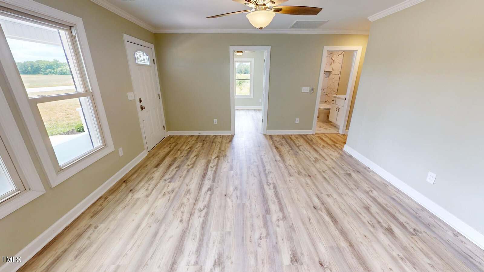 Bright, empty living room with wood flooring, beige walls, white trim, ceiling fan, large windows, front door, and doorways leading to other rooms and a bathroom.