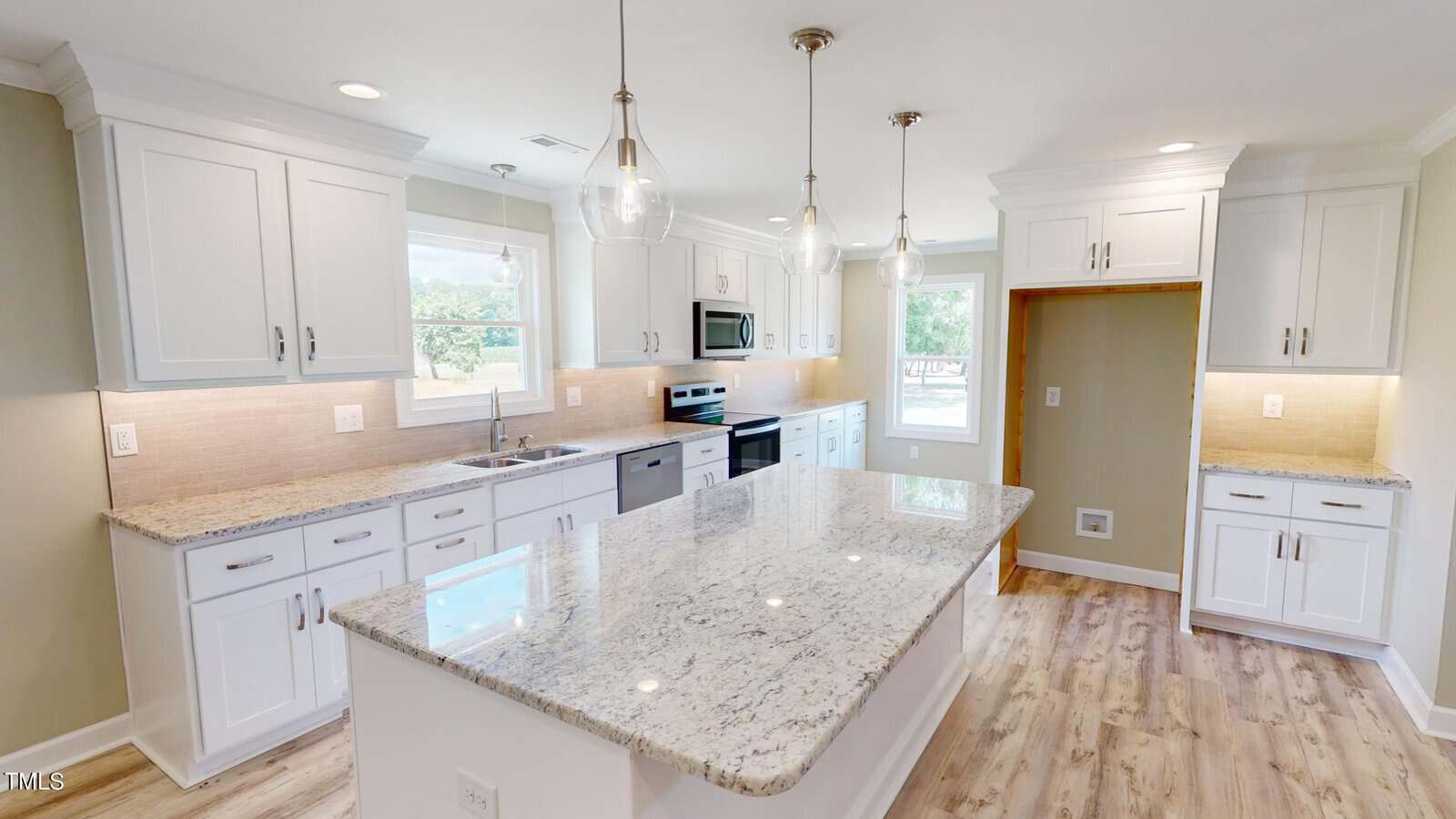 Modern kitchen with white cabinets, granite countertops, stainless steel appliances, glass pendant lights, light wood flooring, and large island with ample workspace. Bright natural light streams through windows.