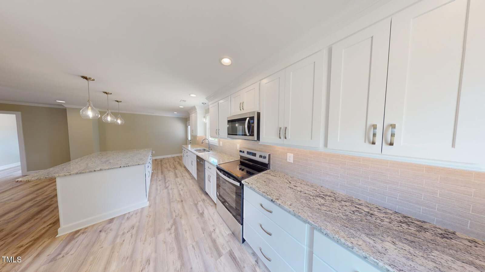 Modern kitchen with white cabinets, stainless steel appliances, granite countertops, a light wood floor, a large island with pendant lights, and a beige tiled backsplash, all brightly lit by recessed and natural lighting.