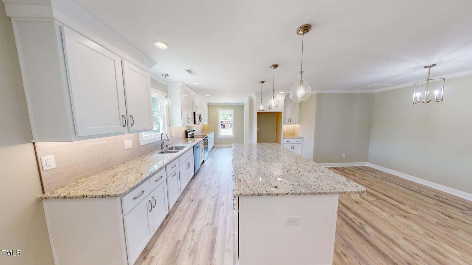 Modern kitchen with white cabinets, granite countertops, a large island, pendant lighting, and light wood floors. Open layout flows into a dining area with a contemporary chandelier. Neutral colors create a bright, spacious feel.