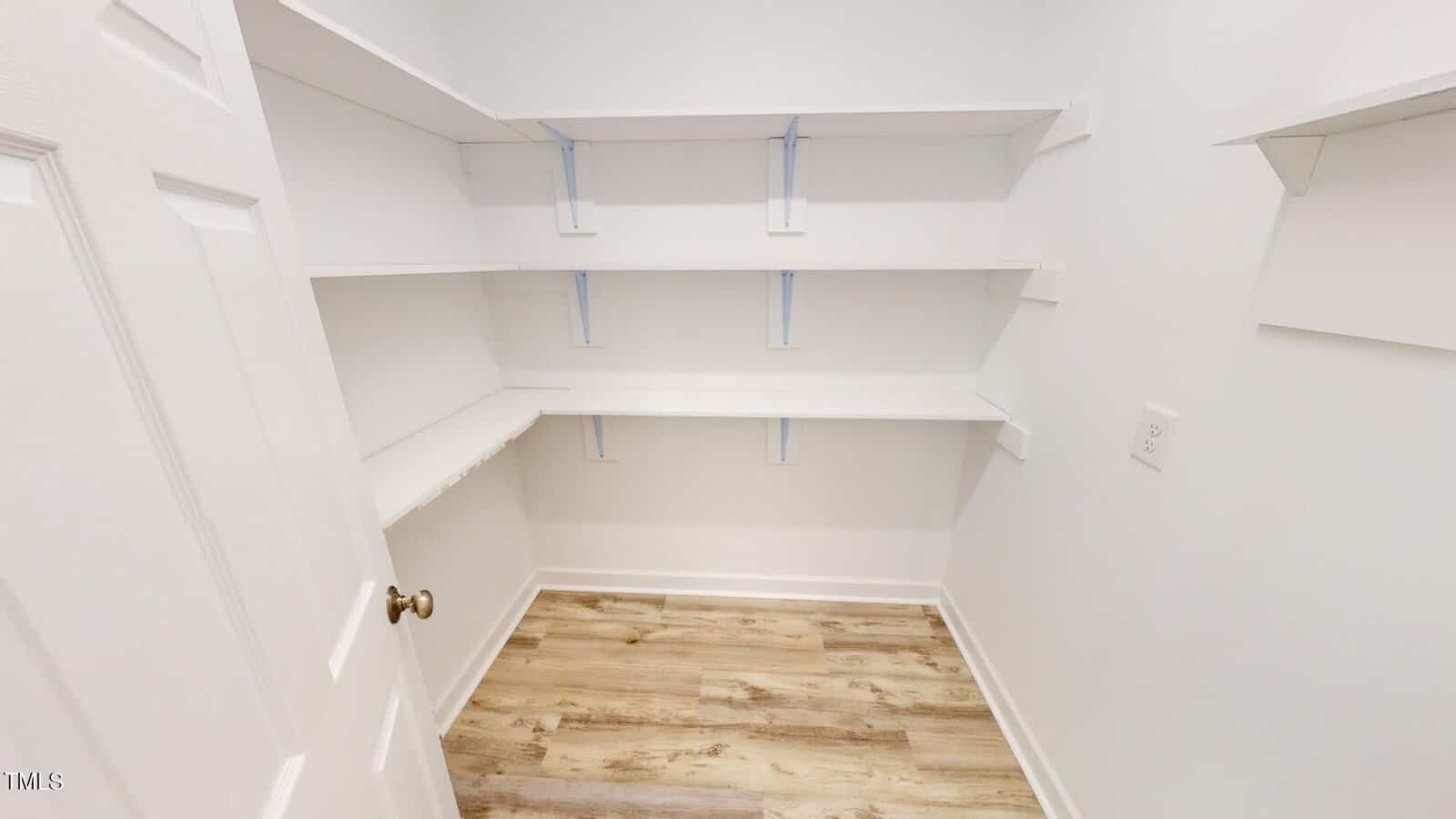 A small, empty walk-in closet with white walls, white shelves on two sides, and a light wood floor. The door is open, and there is a light switch on the right wall.