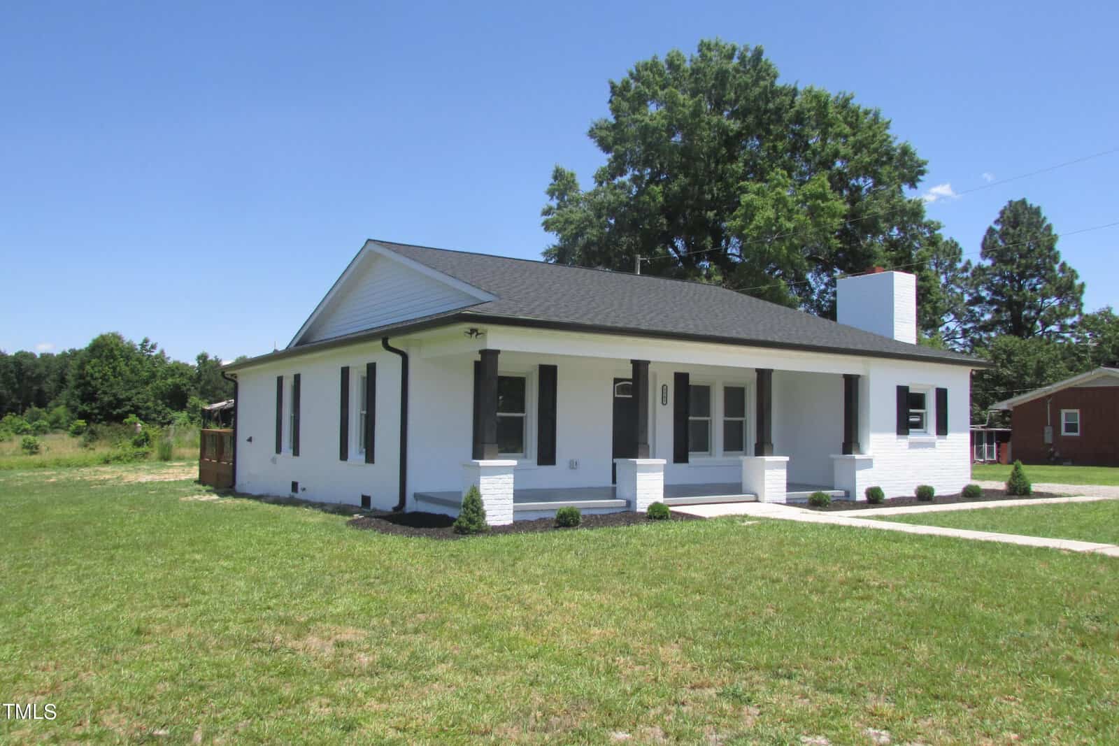 A single-story white brick house with a black roof, a front porch supported by white columns, and large windows, surrounded by grass and a few small shrubs, under a clear blue sky.