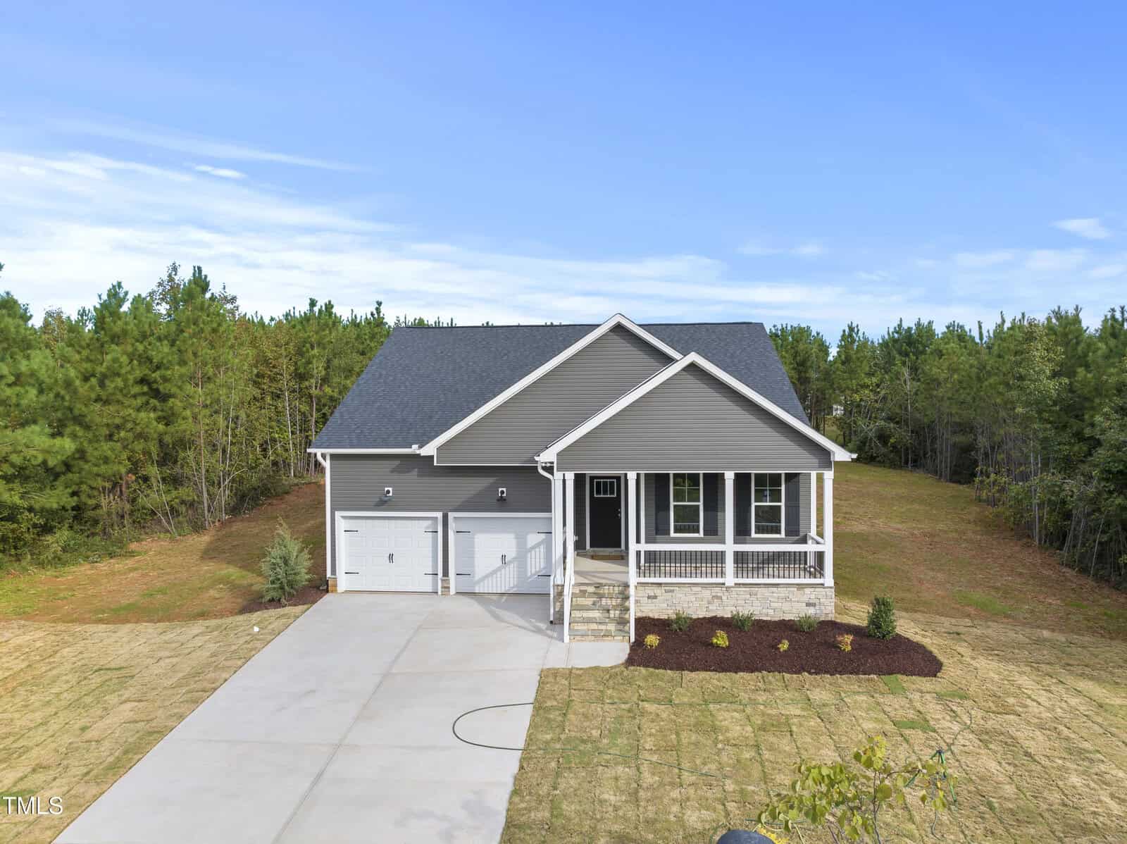 A modern gray single-story house with a covered front porch, two-car garage, and a wide driveway, surrounded by a yard and backed by dense green trees under a blue sky.