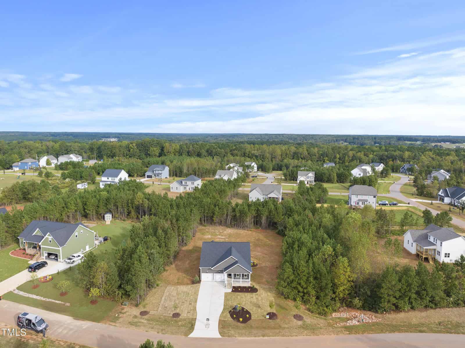 Aerial view of a suburban neighborhood with houses surrounded by trees and greenery under a blue sky. A central house with a driveway is in the foreground, bordered by wooded areas and open lawns.