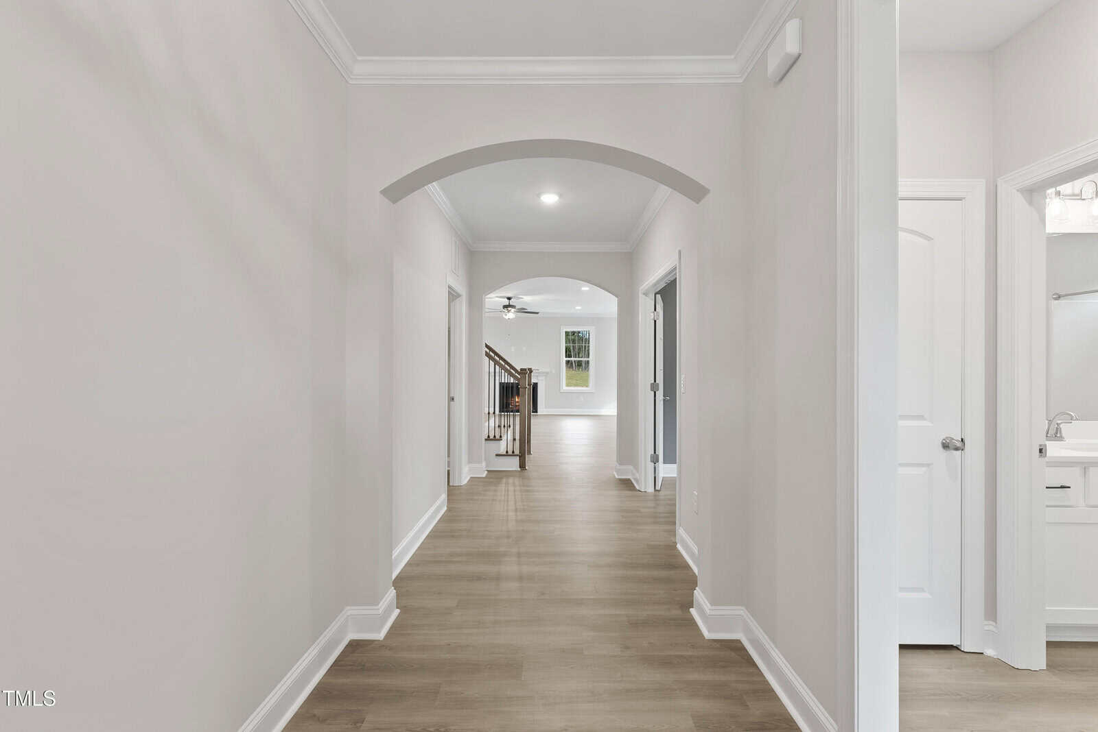 A bright hallway with light wood flooring, arched entryway, white walls, and crown molding, leading to a staircase and living area; a bathroom is visible on the right.