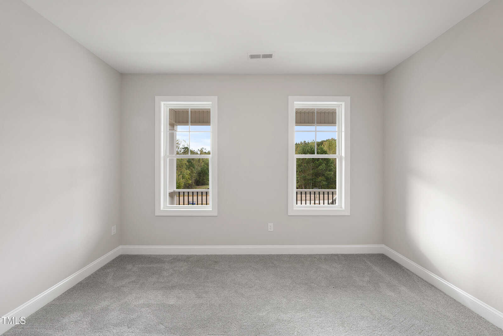 Empty room with light gray walls, two white-trimmed windows, and gray carpet. The windows show a view of greenery and blue sky outside. The room appears clean and well-lit with natural light.