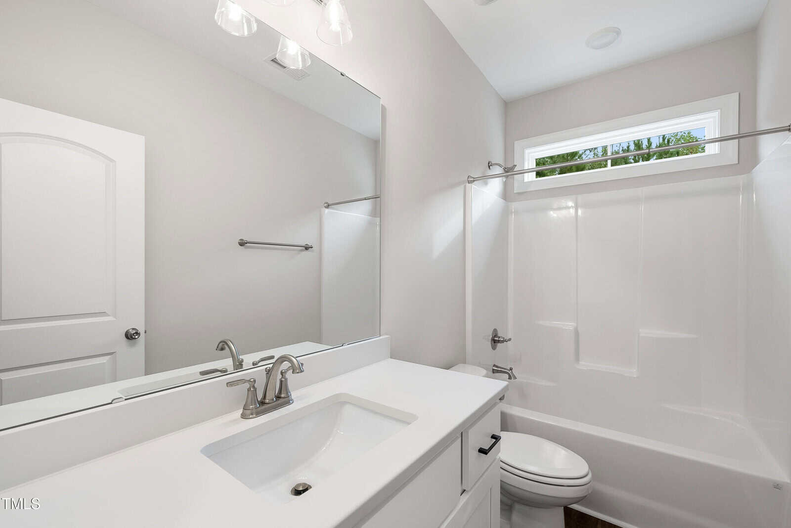 Modern, clean bathroom with white walls and fixtures, a large mirror, sink with silver faucet, toilet, and a bathtub-shower combo under a rectangular window. Natural light brightens the space.