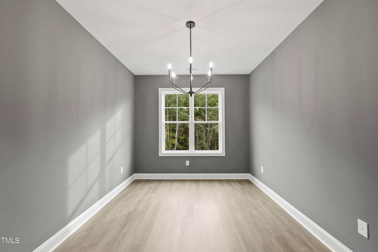 A modern, empty room with gray walls, light wood flooring, a window showing green trees outside, and a contemporary black chandelier hanging from the ceiling.